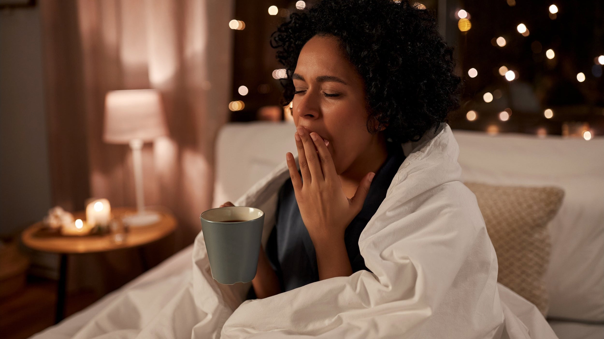 A woman yawns in bed with a cup of coffee. Learn the 10-3-2-1-0 sleep rule: stop caffeine 10 hours before bed, food 3 hours, work 2 hours, screens 1 hour, and hit snooze 0 times for better sleep.