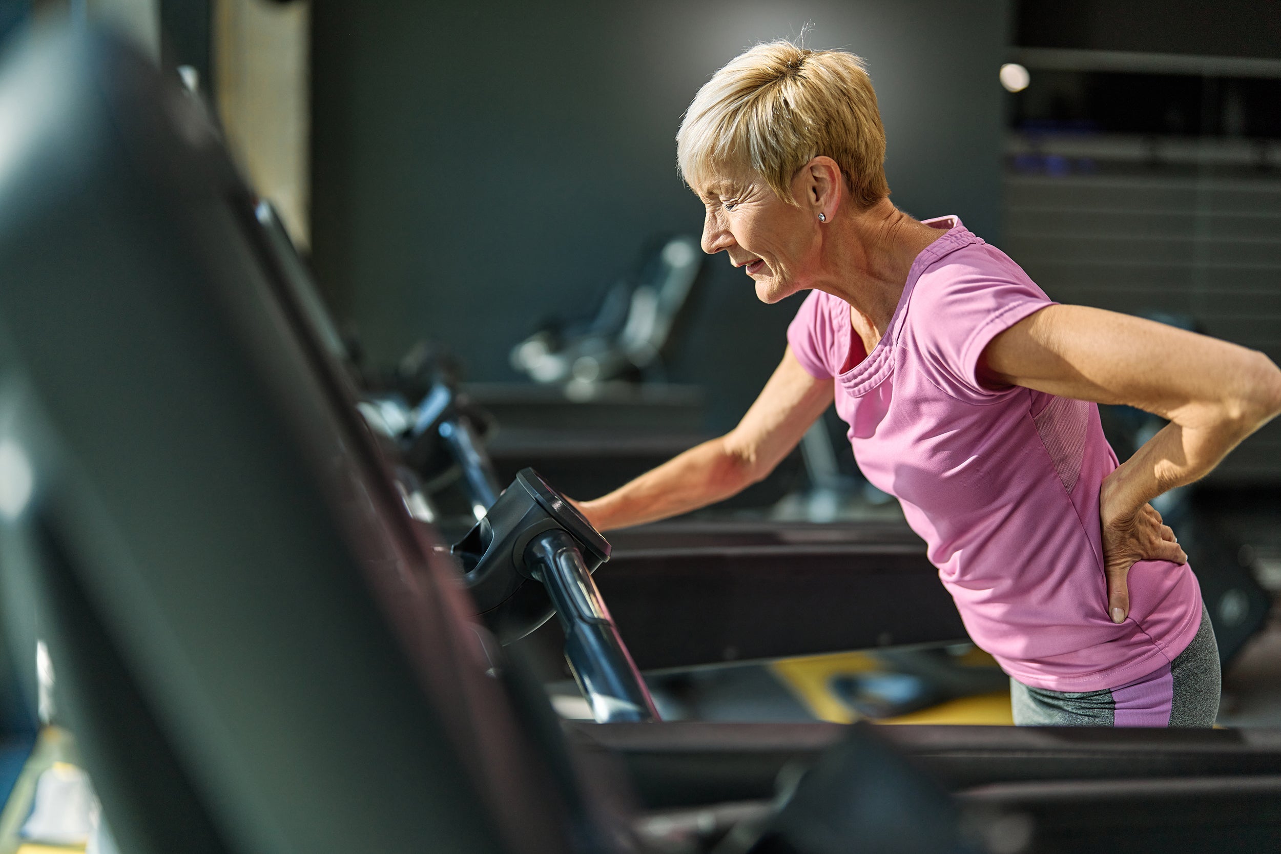 A woman clutches her back while walking on the treadmill. If you're suffering from back pain, check out our comprehensive guide on finding the best pillow for back pain relief.