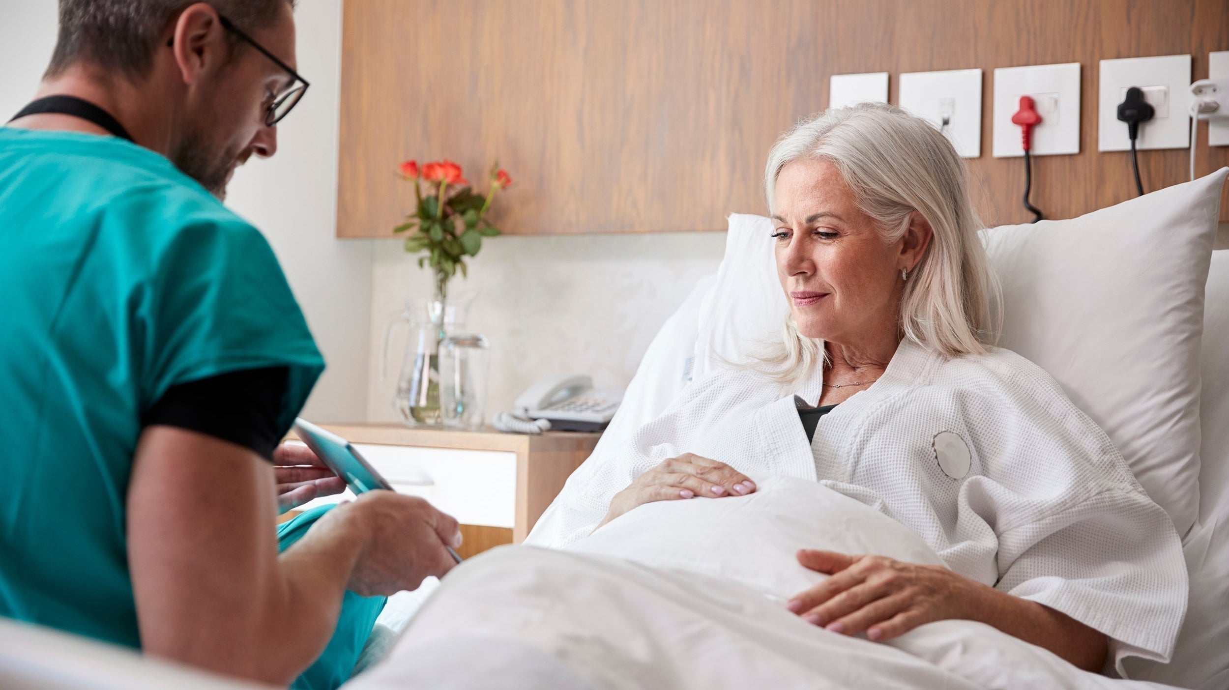 A woman talks to a doctor in a hospital bed. If you're preparing for a tummy tuck, how to sleep after the surgery can make a huge difference in overall healing. Learn more in our complete guide.