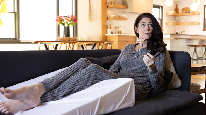 A woman rests on the couch using a single orthopedic wedge pillow for legs from the Sleep Again Pillow System.