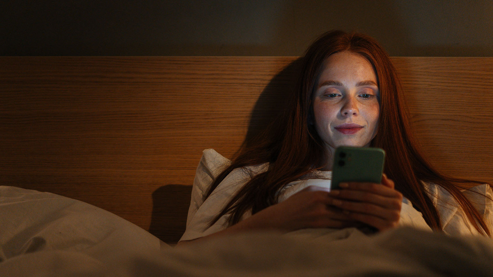 A woman reading her phone in bed. Learn everything you need to know about breast fat transfer recovery, including sleep - one of the most critical recovery pieces.