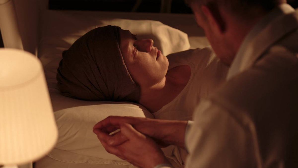 Person resting on their back in bed while a caregiver holds their hand, with a lamp and glass of water on the nightstand.