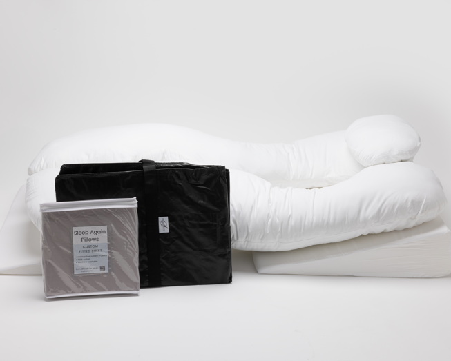 A large white body pillow and wedge pillow are arranged on a white background. In front, a black carrying bag and a packaged Sleep Again Pillows pillowcase are displayed.