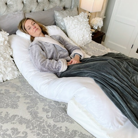 A woman in a gray robe is lying on a bed, resting on a large white body pillow and covered with a gray blanket in a well-decorated bedroom.