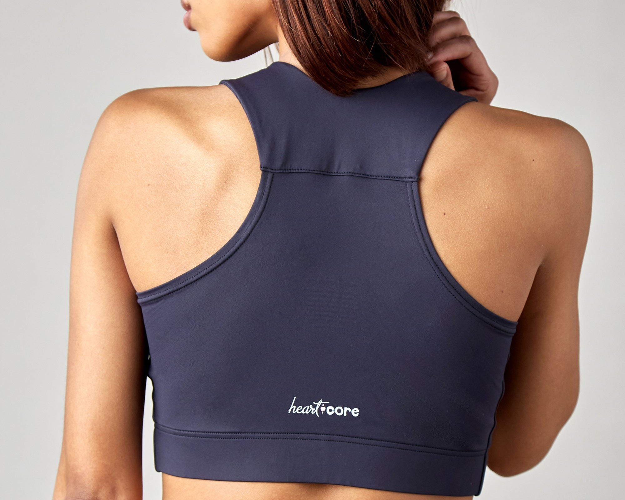 A woman with straight brown hair is shown from the back, wearing a navy blue racerback sports bra with the heartcore logo printed in white on the lower back.