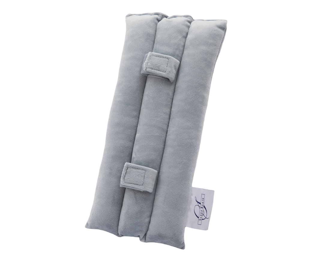 The Seat Belt Pillow is a light gray, rectangular padded fabric cushion designed for chest support. It features three cushioned sections, two rectangular fabric loops, and a small white logo tag on the side. The background is plain white.