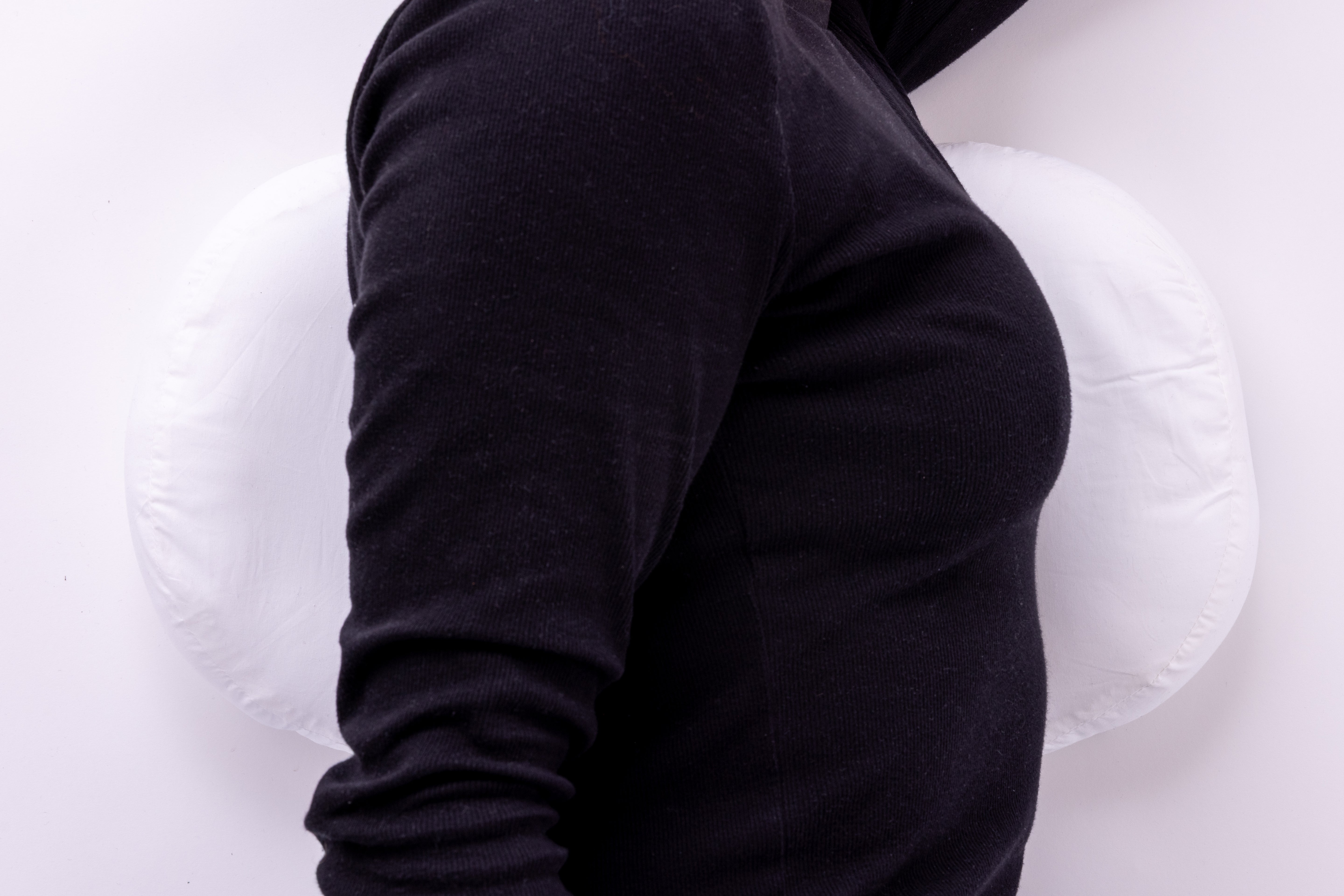 A person in a black long-sleeve shirt sits with their back against the Side Sleeping Chest Pillow, which supports their lower and middle back. The background is plain and light-colored.