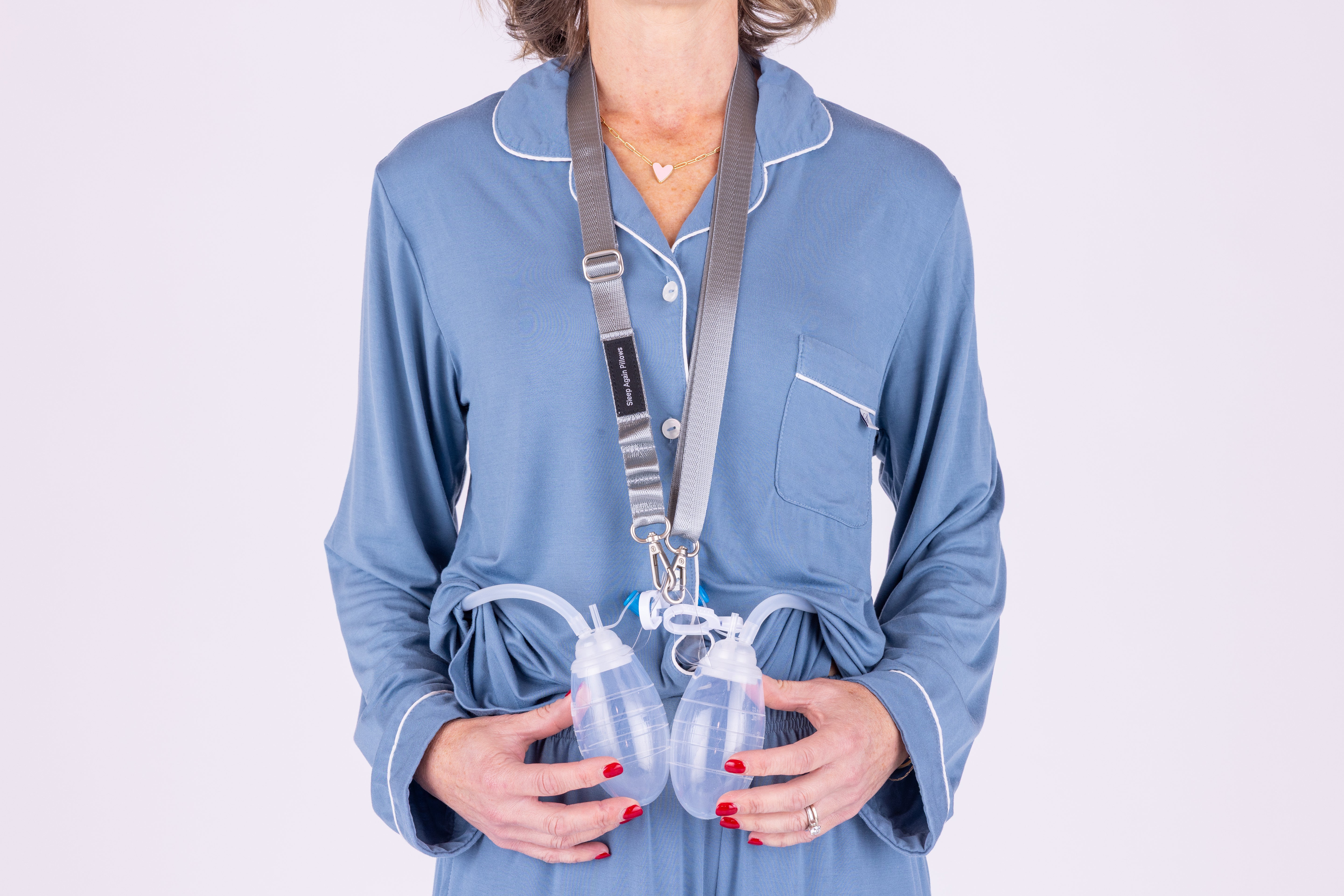 A woman in blue pajamas uses the Sleep Again Surgical Drain Holder lanyard to carry two JP surgical drains around her neck, displaying the drains and tubing. Her face is not visible.