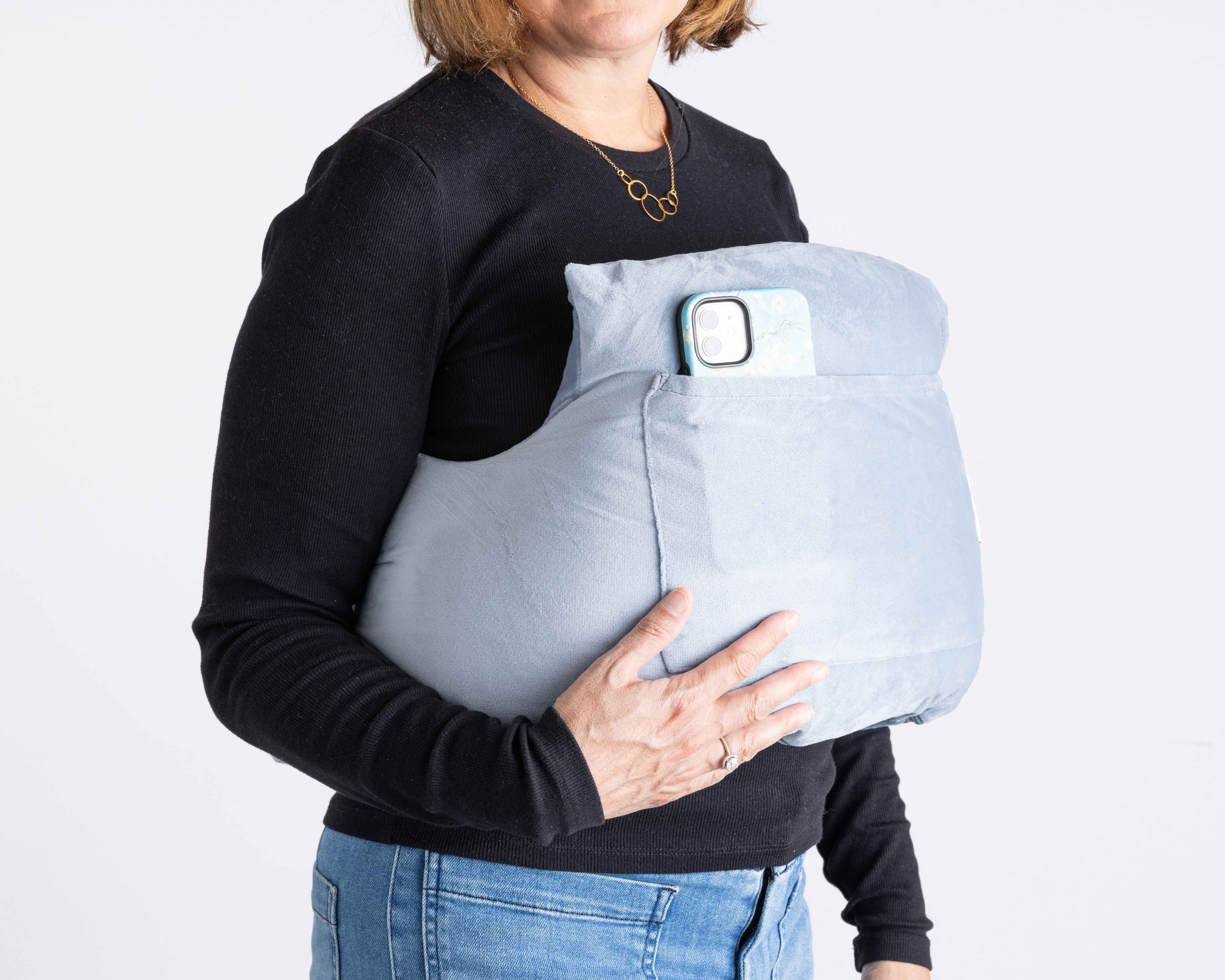 A woman in a black long-sleeve shirt and jeans holds the Chest Pillow, a large light blue post-surgery pillow with an external pocket that holds a smartphone.