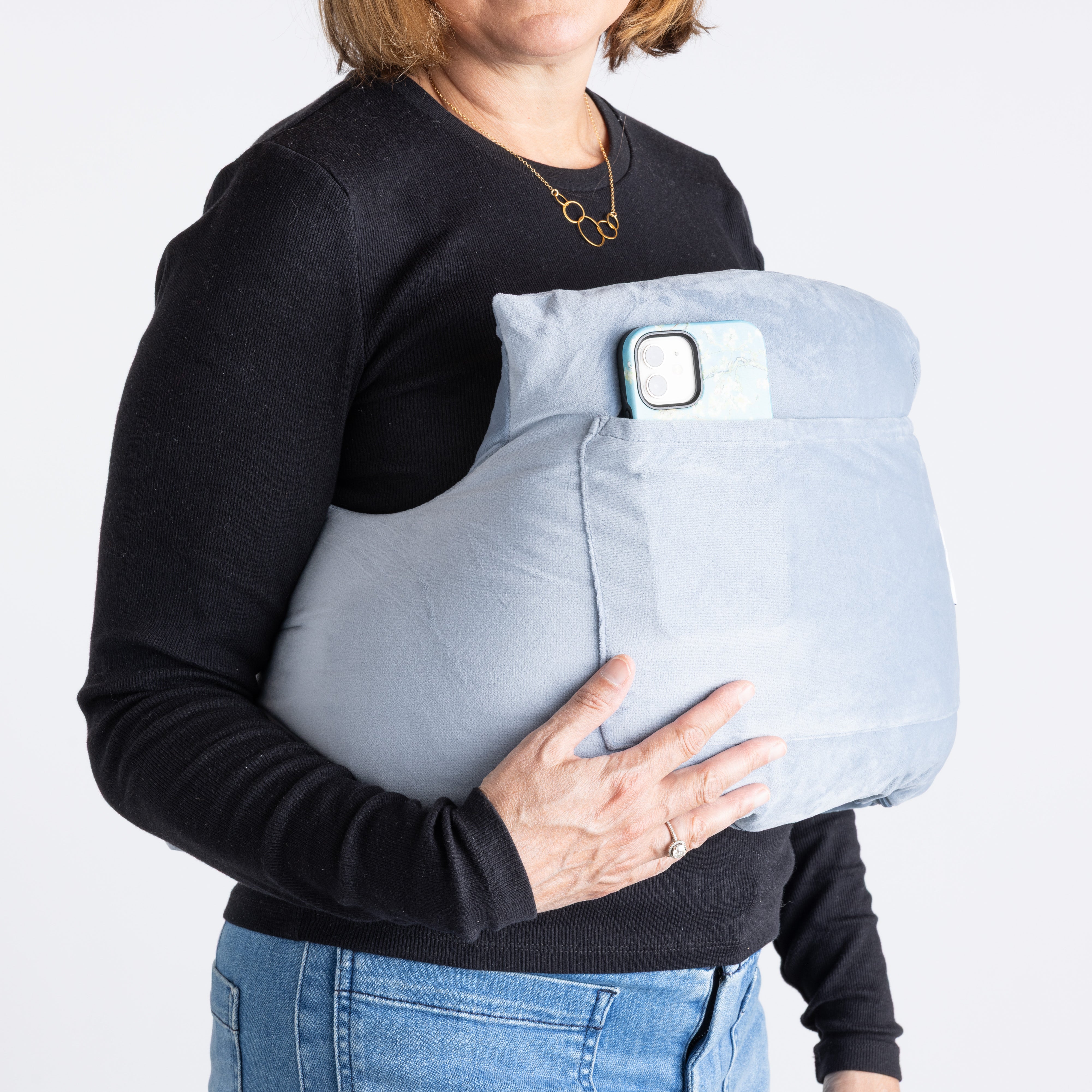 A woman in a black long-sleeve shirt and jeans holds the Chest Pillow, a large light blue post-surgery pillow with an external pocket that holds a smartphone.