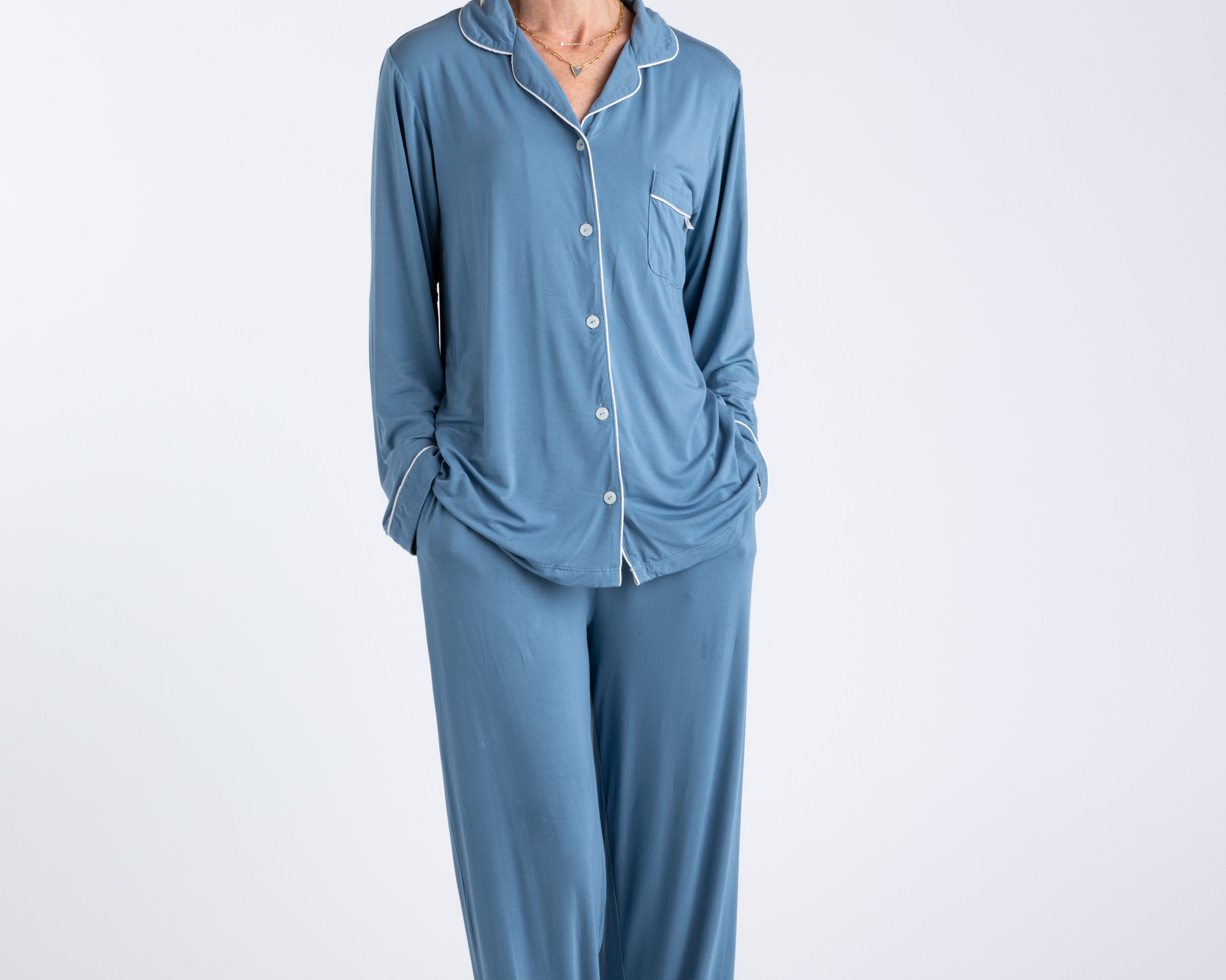 Blonde woman with glasses wearing blue bamboo pajamas with contrasting white piping standing on white background with her hands in her pockets.