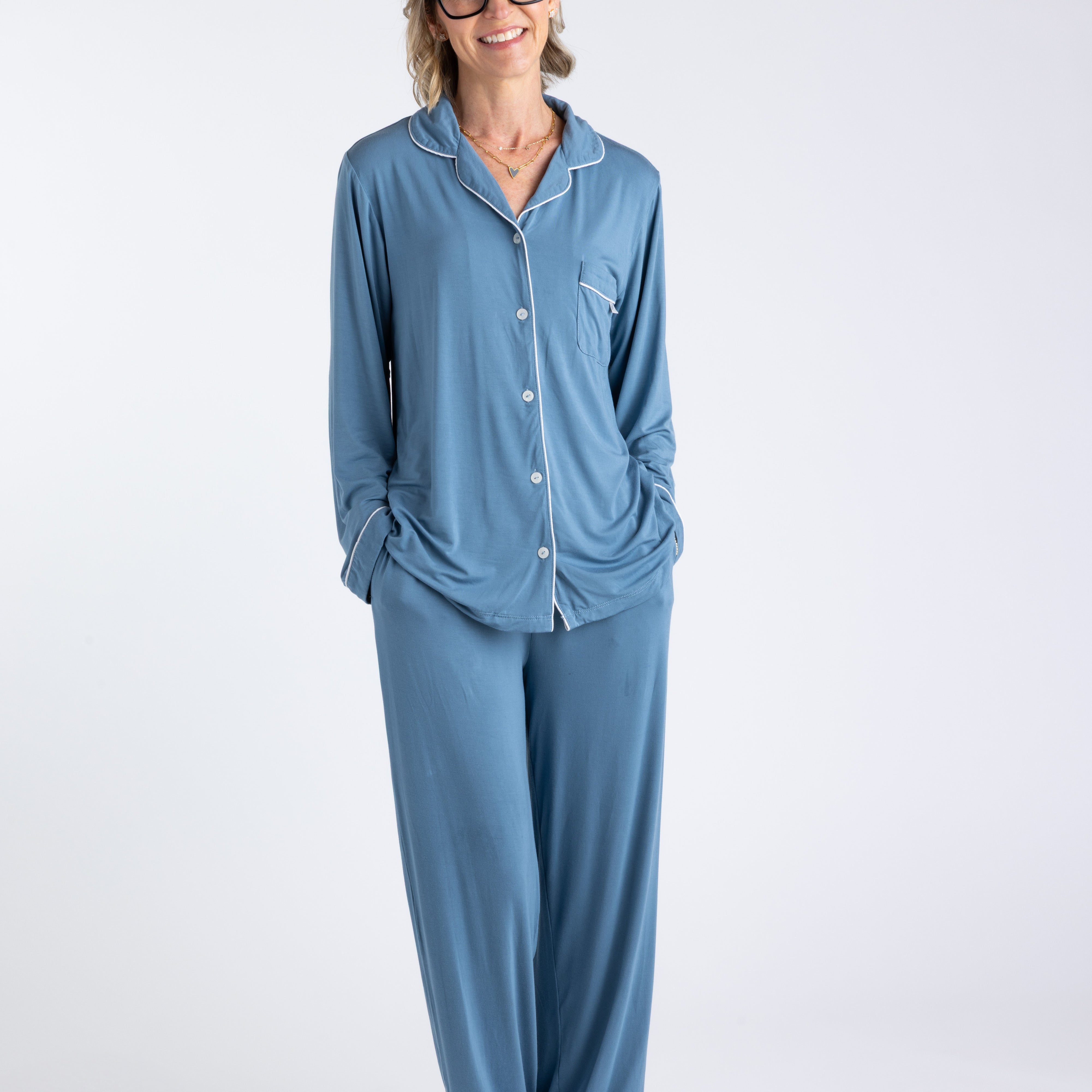Blonde woman with glasses wearing blue bamboo pajamas with contrasting white piping standing on white background with her hands in her pockets.