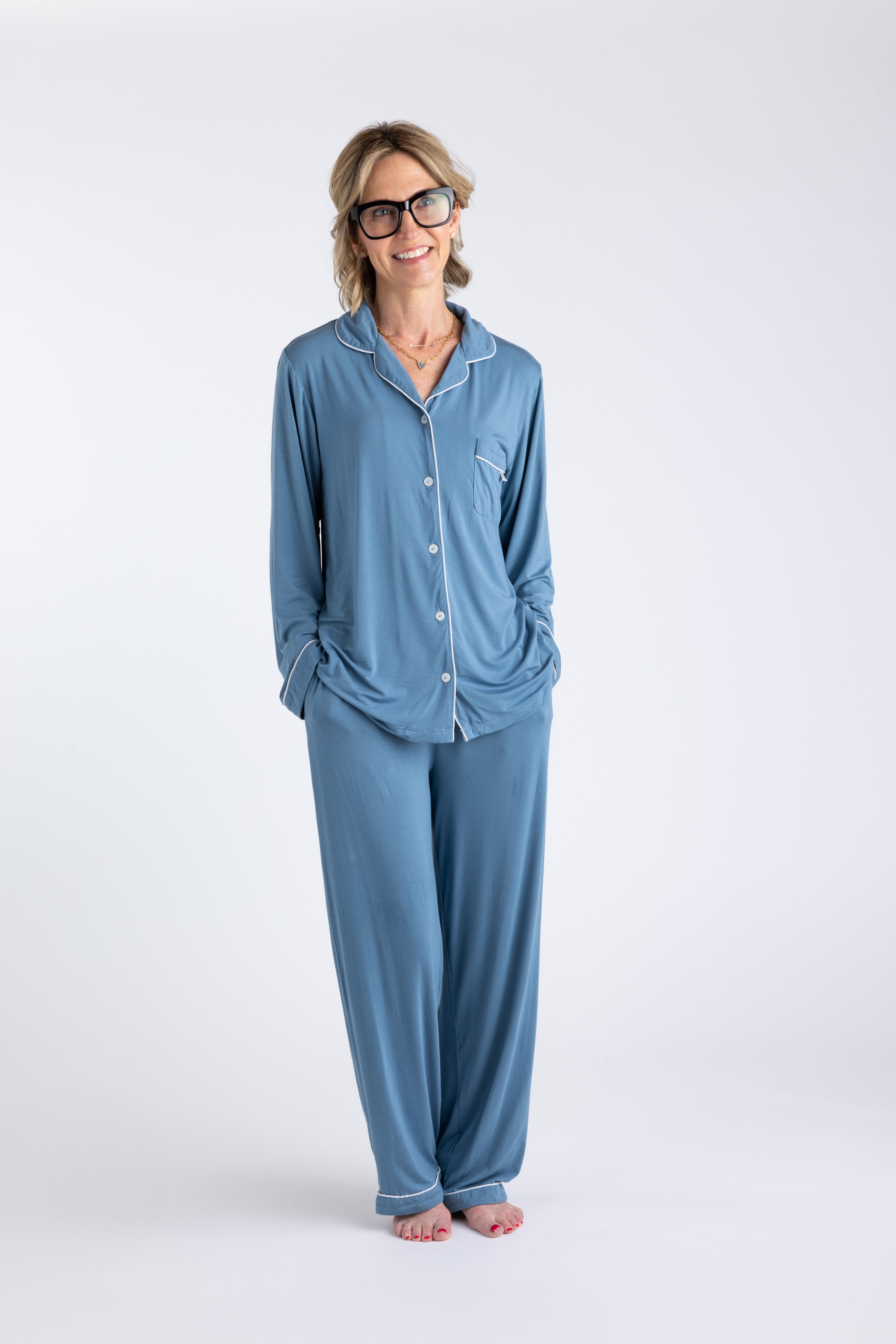 Blonde woman with glasses wearing blue bamboo pajamas with contrasting white piping standing on white background with her hands in her pockets.