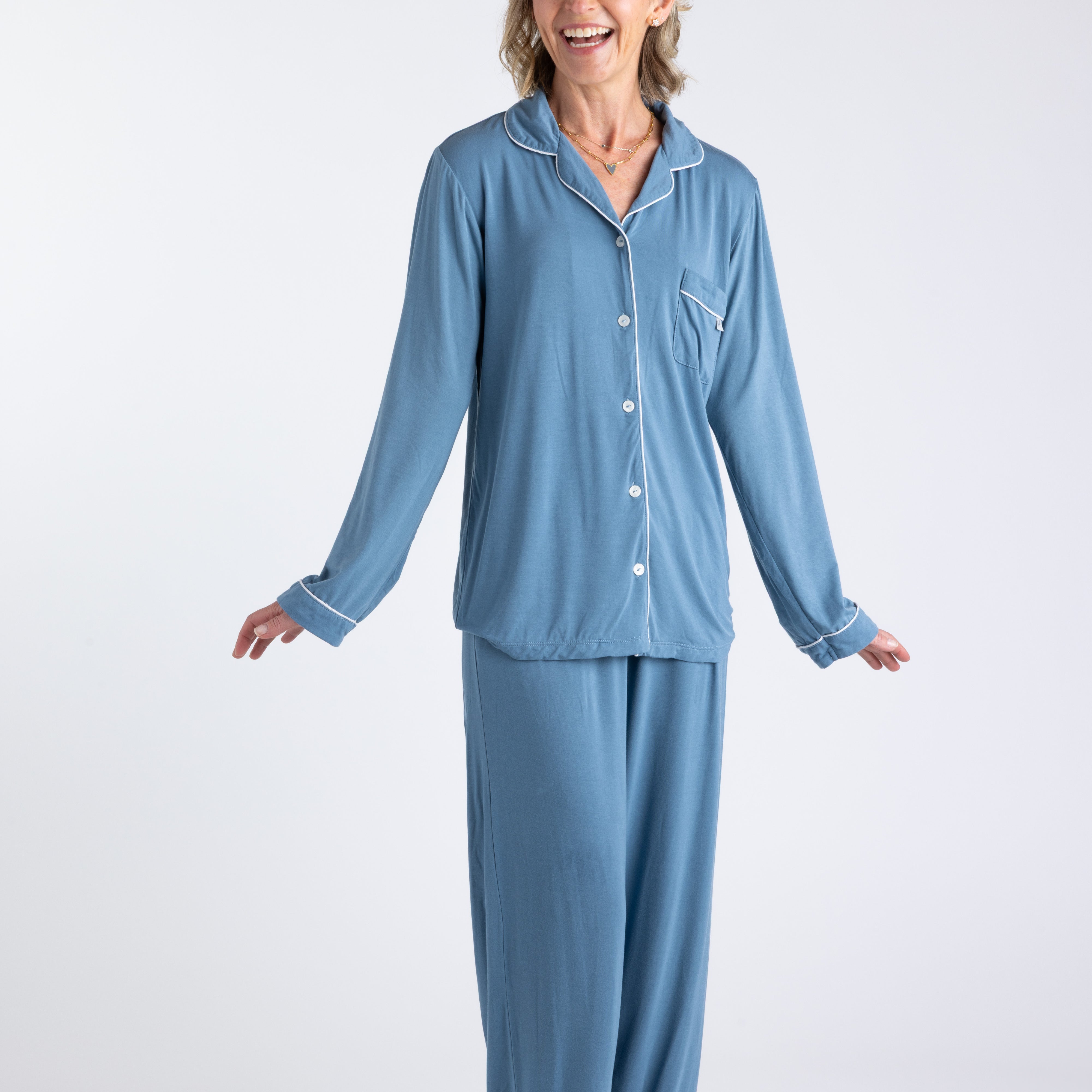 Blonde woman standing smiling wearing blue bamboo pajamas with front buttons and white piping