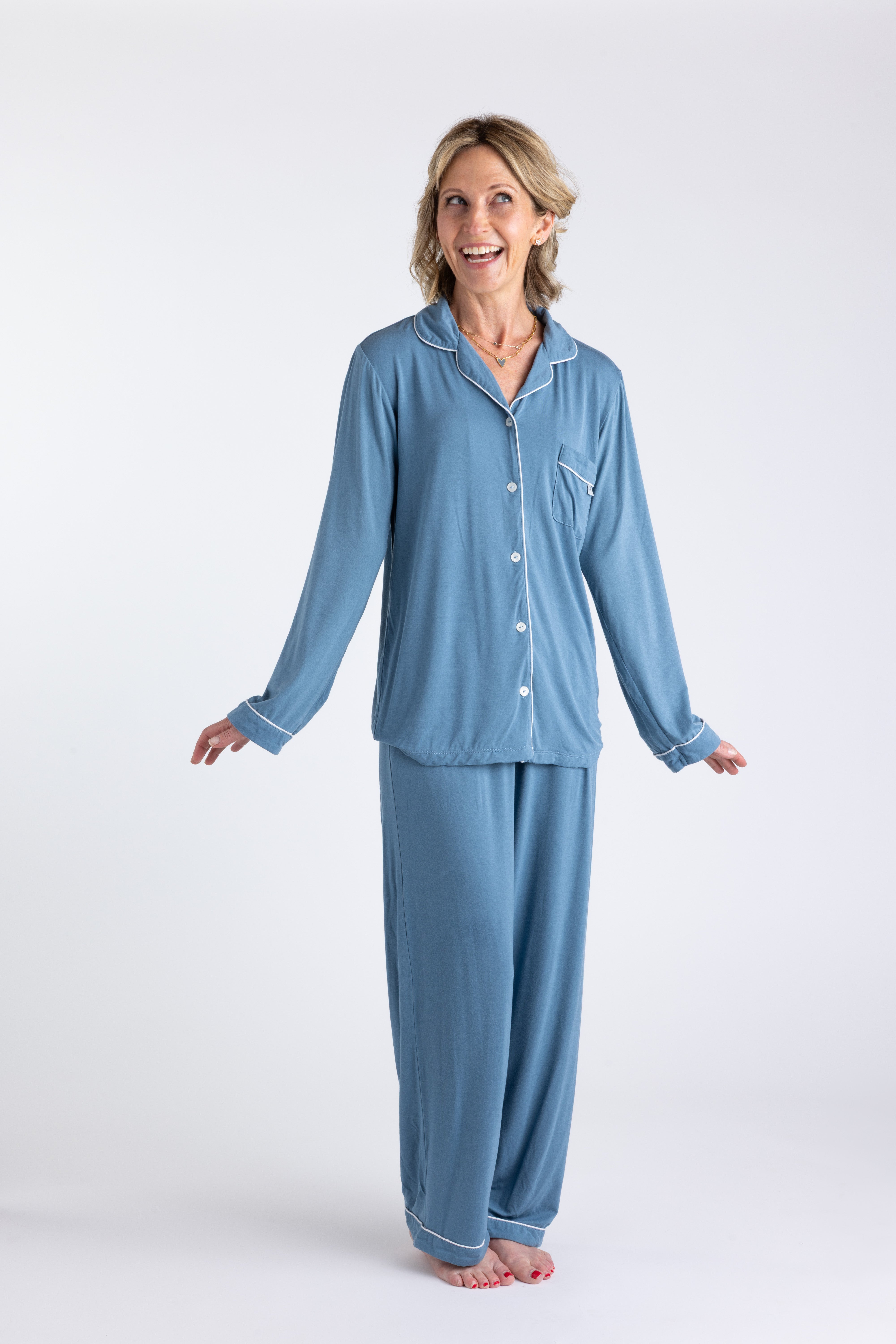 Blonde woman standing smiling wearing blue bamboo pajamas with front buttons and white piping