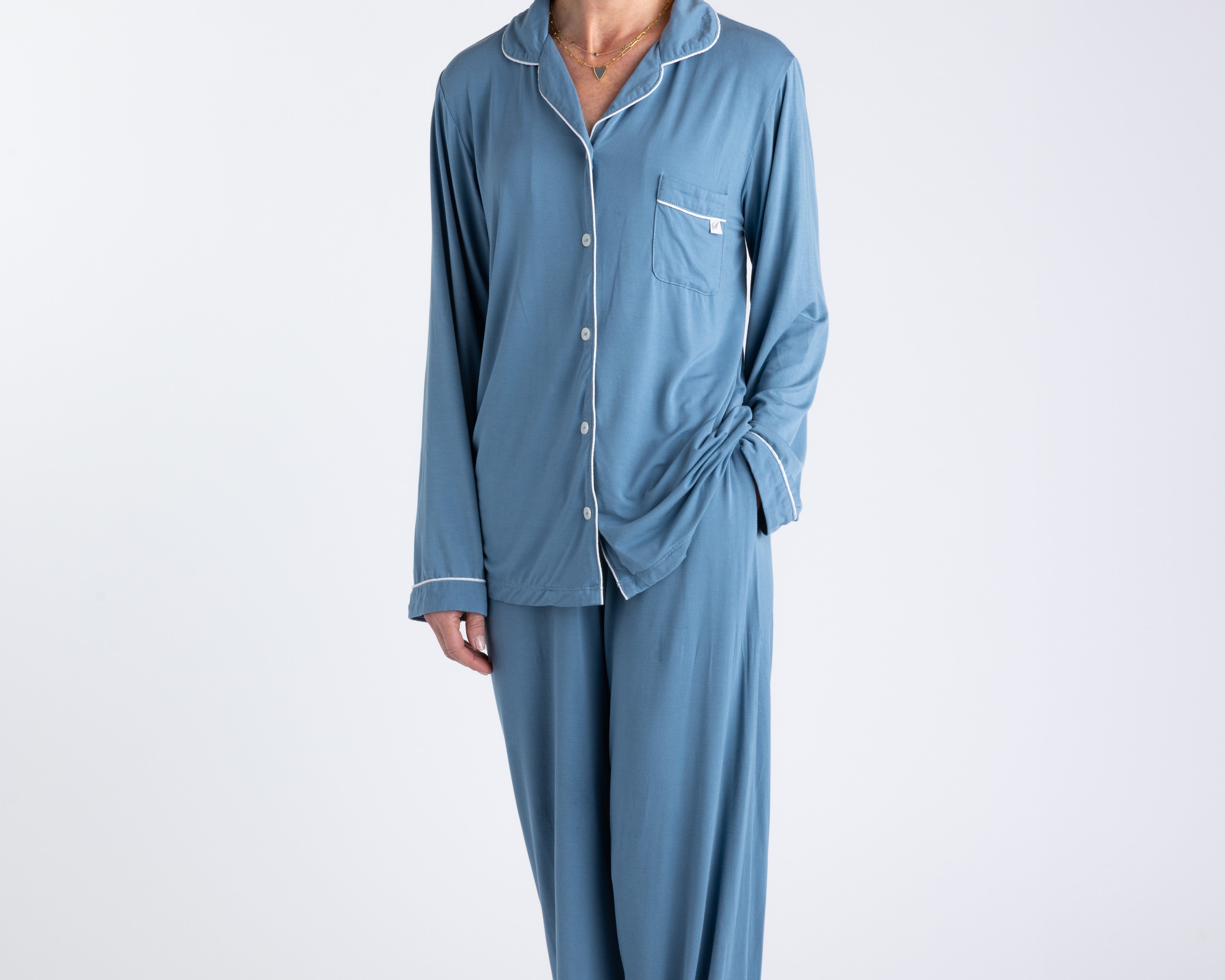 A woman standing and wearing a long sleeve blue bamboo pajama set with contrast piping and button-up shirt style. The set includes pants with an elastic waistband.