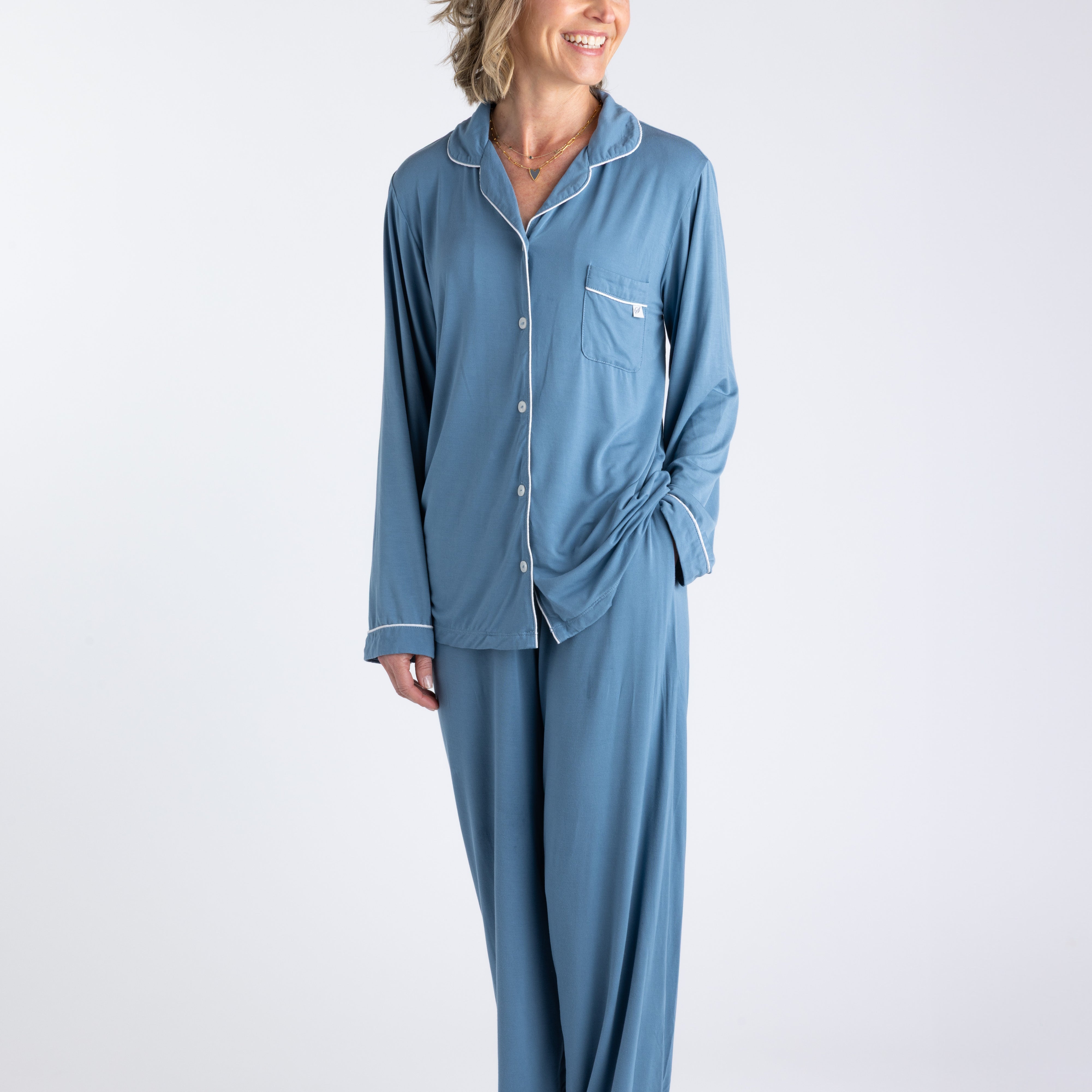 A woman standing and wearing a long sleeve blue bamboo pajama set with contrast piping and button-up shirt style. The set includes pants with an elastic waistband.