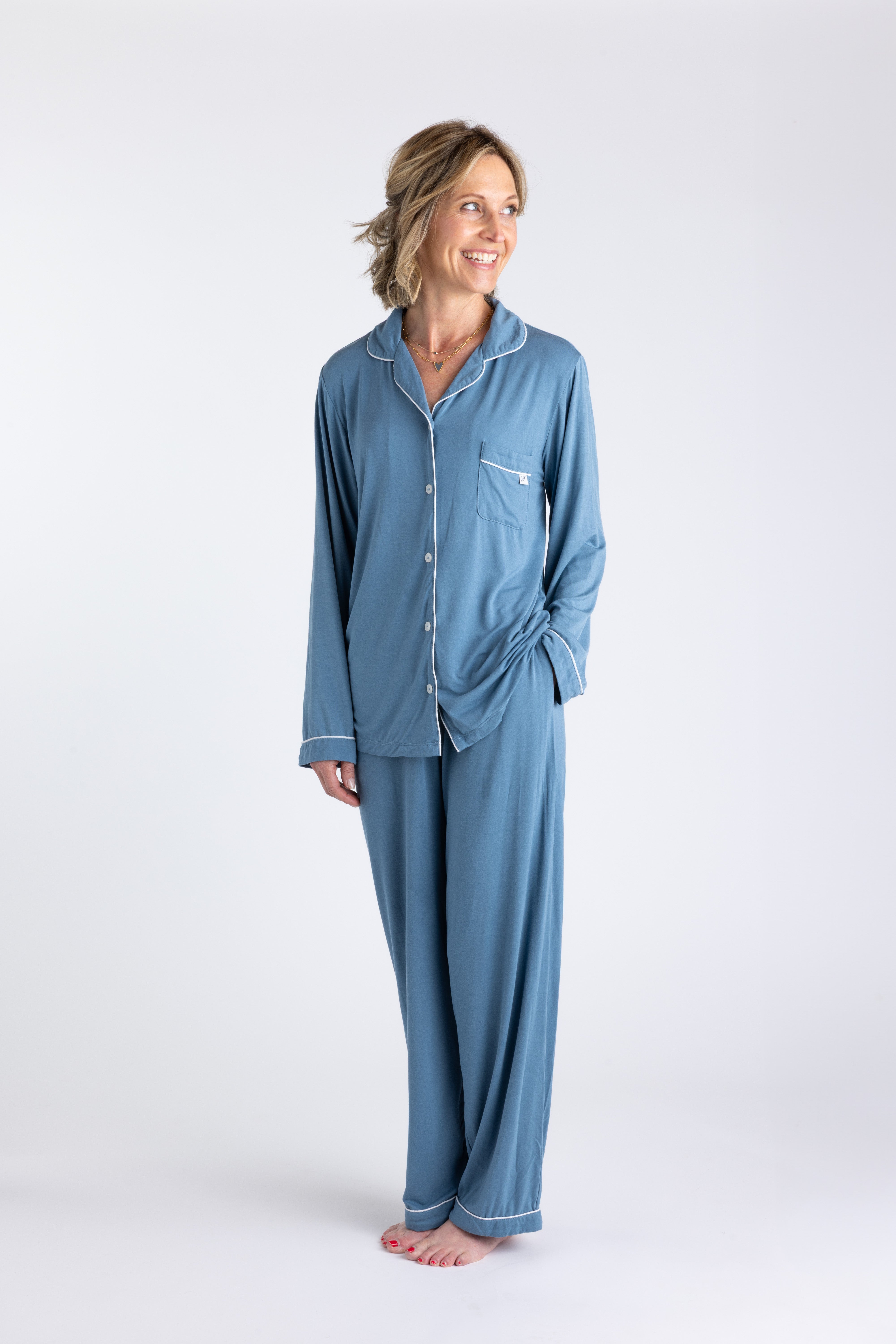 A woman standing and wearing a long sleeve blue bamboo pajama set with contrast piping and button-up shirt style. The set includes pants with an elastic waistband.