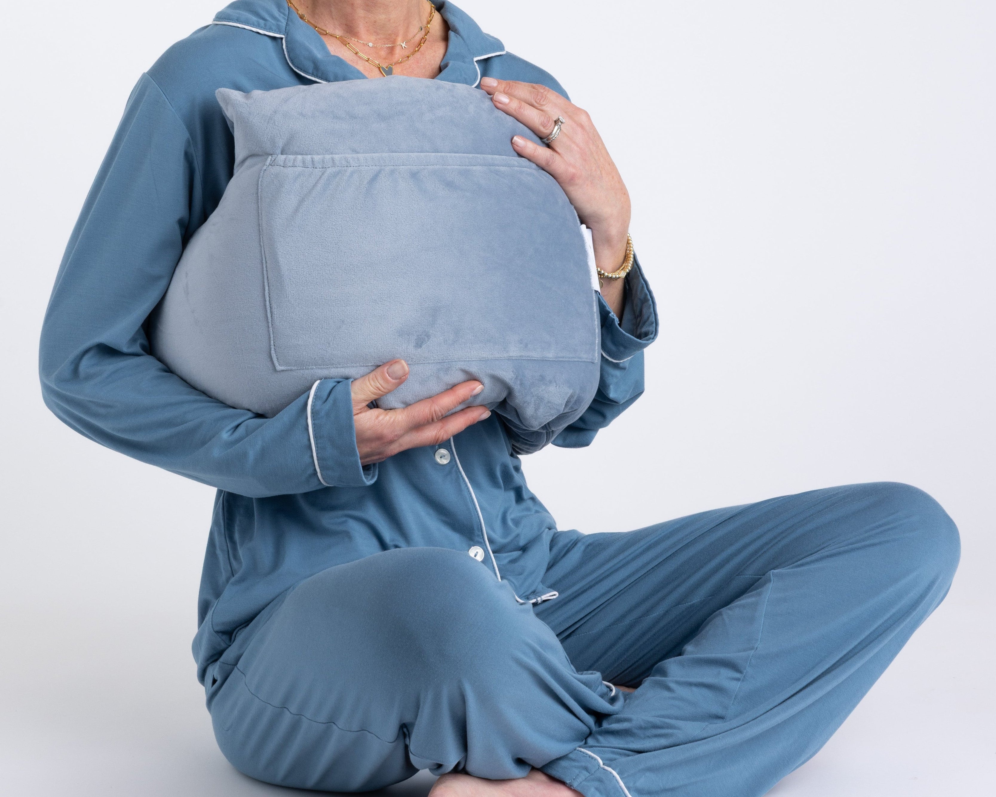 A person in blue pajamas sits cross-legged, hugging the Chest Pillow—a large, light gray pillow—to their chest with both hands. The background is plain white, and the person's head is not shown.