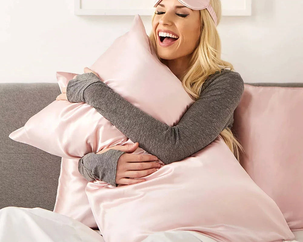 A woman wearing a Mulberry Silk Sleep Eye Mask with Silk Covered Elastic Strap and a gray long-sleeve top smiles while hugging a shiny pink satin pillow, sitting on a bed with matching satin pillows and white bedding.