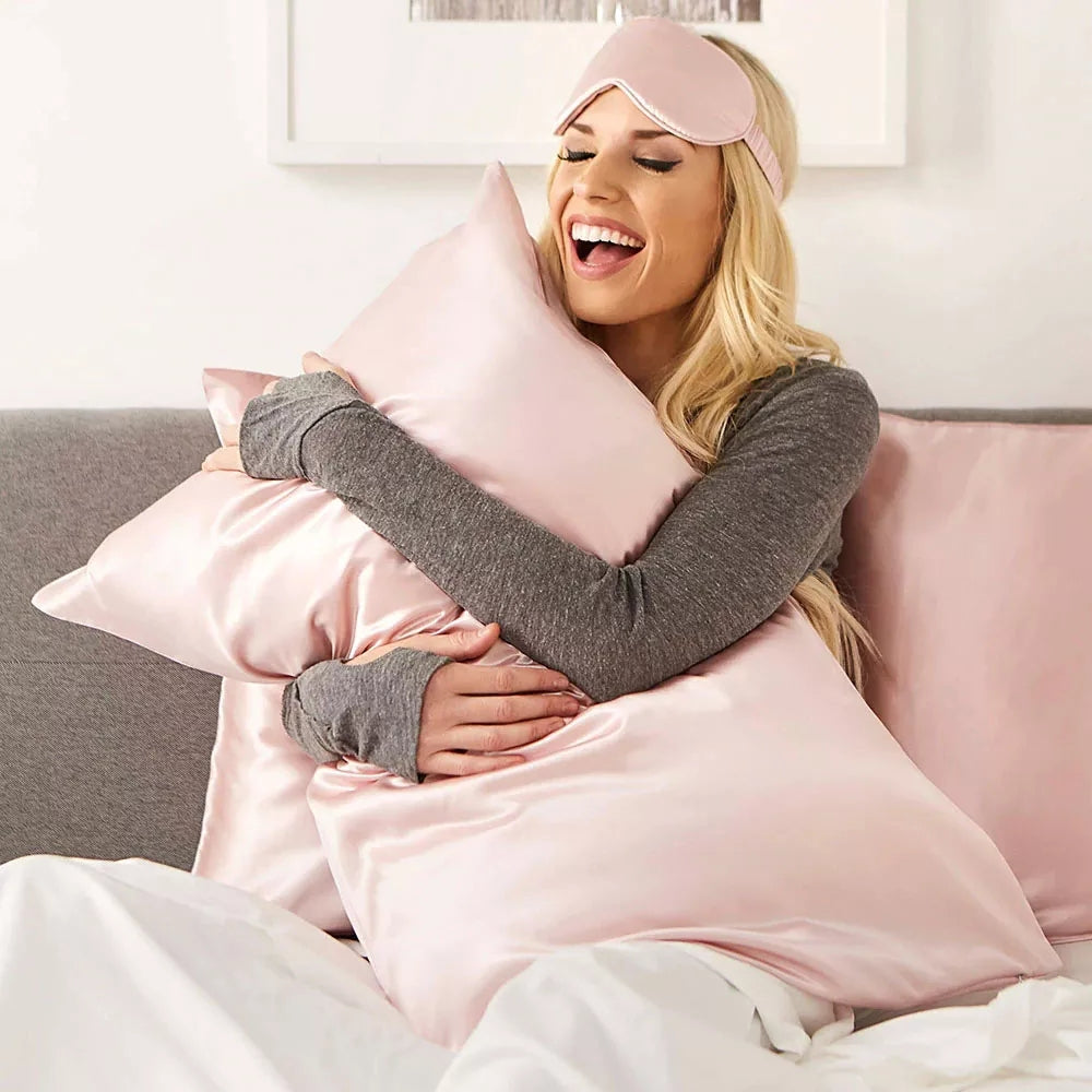 A woman wearing a Mulberry Silk Sleep Eye Mask with Silk Covered Elastic Strap and a gray long-sleeve top smiles while hugging a shiny pink satin pillow, sitting on a bed with matching satin pillows and white bedding.