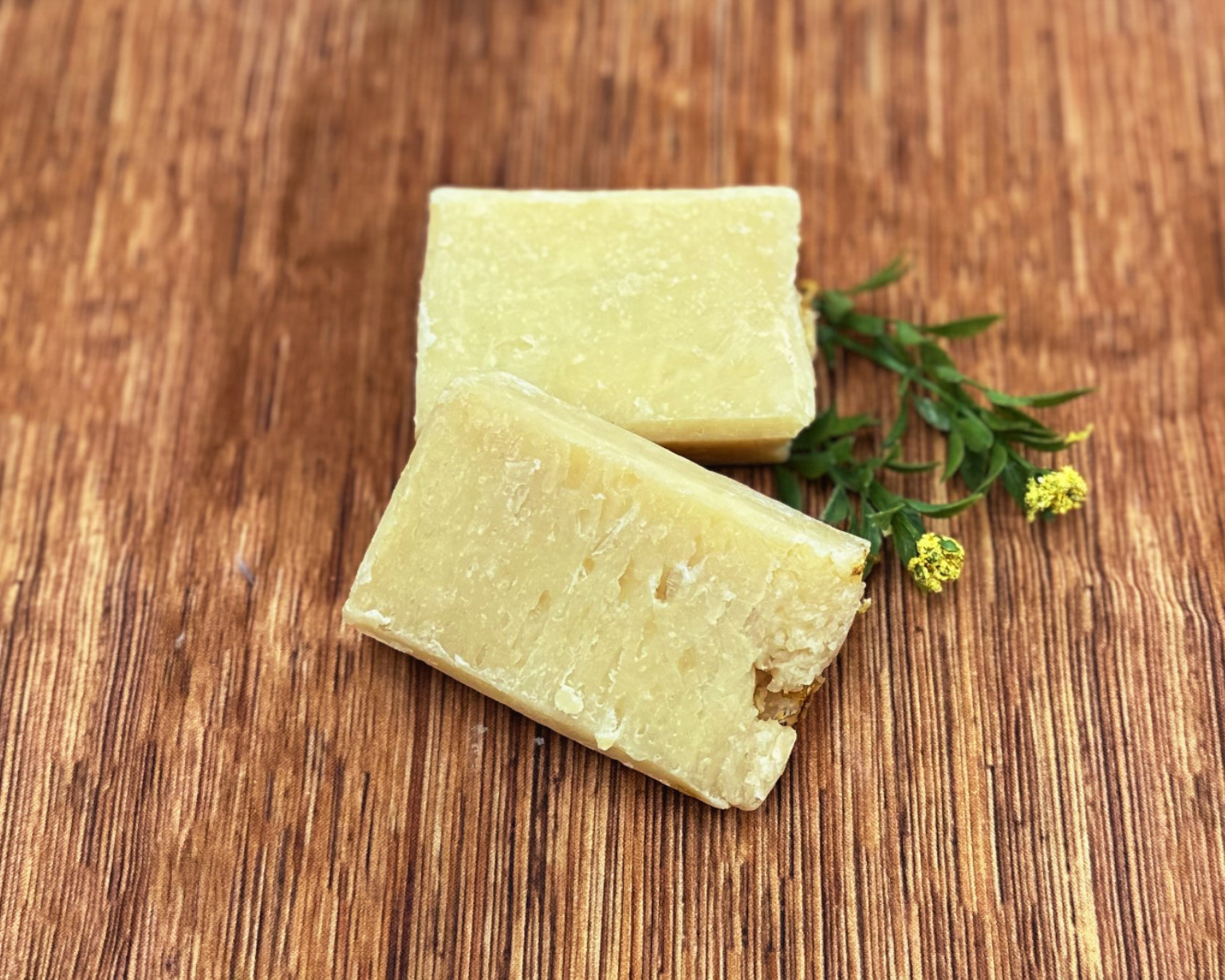 Two blocks of pale yellow Premium Soothing Skin & Lips rest on a wooden surface, adorned with a green sprig and tiny yellow flowers—an ideal addition to a soothing cancer care gift box.