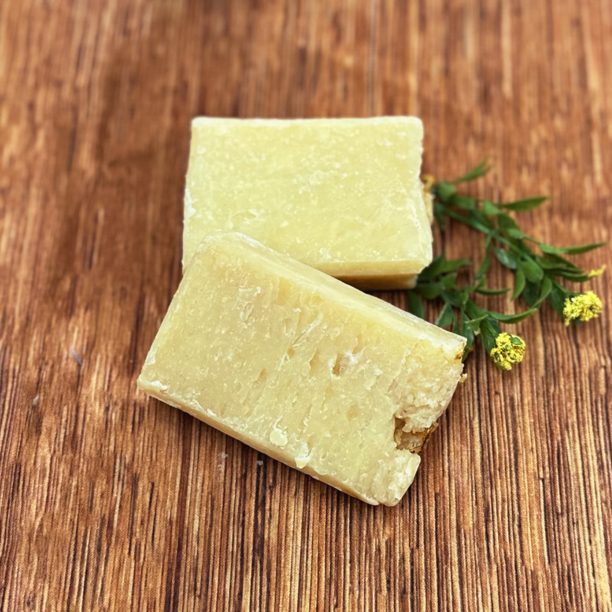 Two blocks of pale yellow Premium Soothing Skin & Lips rest on a wooden surface, adorned with a green sprig and tiny yellow flowers—an ideal addition to a soothing cancer care gift box.