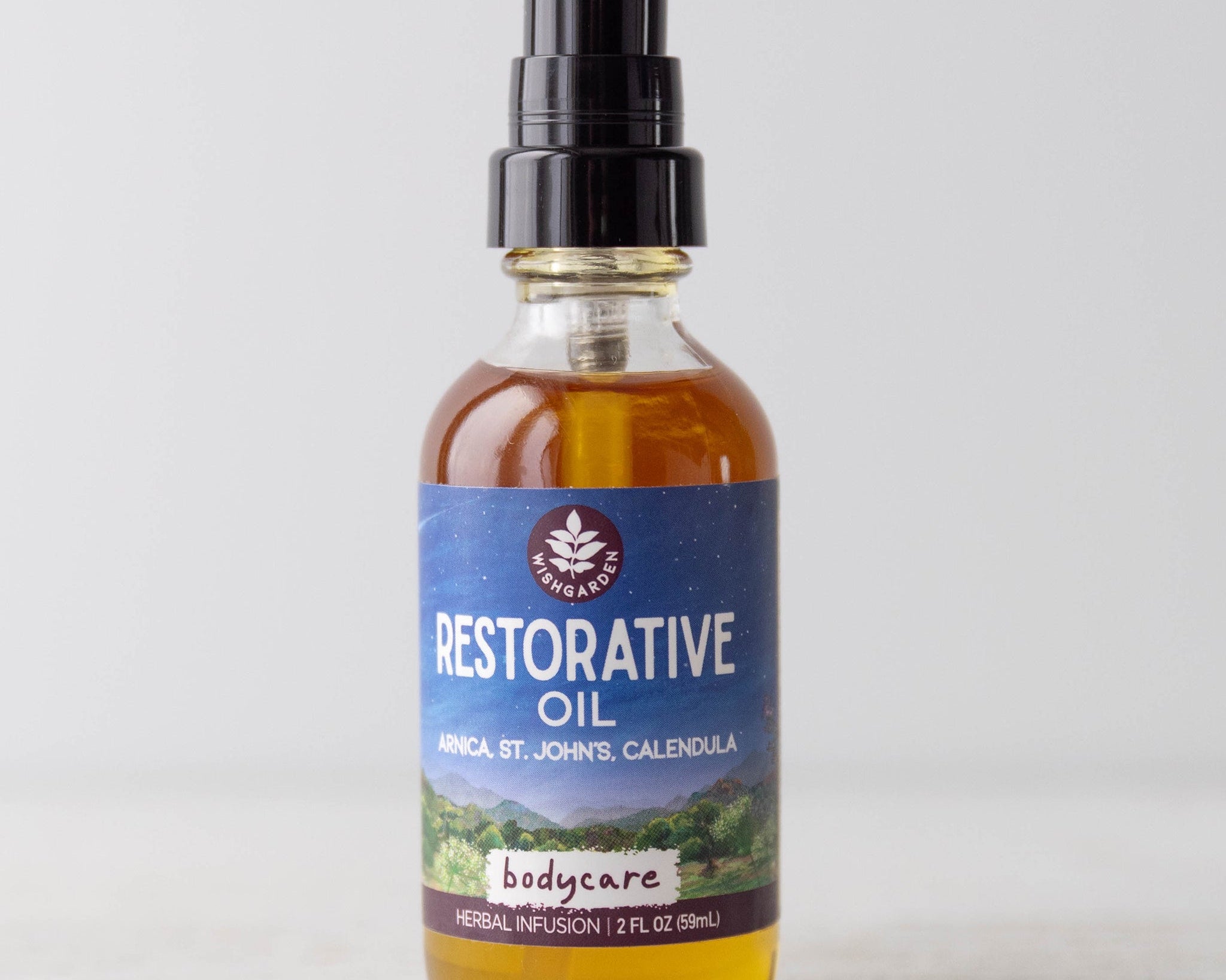 A clear glass bottle with a black pump, labeled Restorative Oil - Arnica, St. John's Wort & Calendula, sits on a light surface. The herb-infused body oil is amber in color and has a purple-blue label with white text.