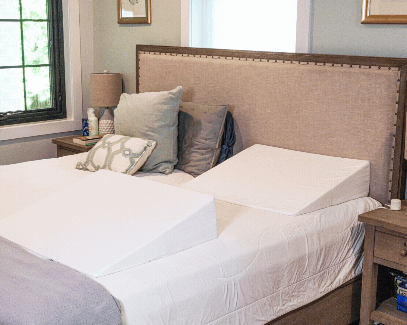 A neatly made bed with a beige headboard features two white BBL Add On Set foam wedge pillows side by side. Decorative pillows rest against the headboard, and matching lamps sit on wooden nightstands on either side.