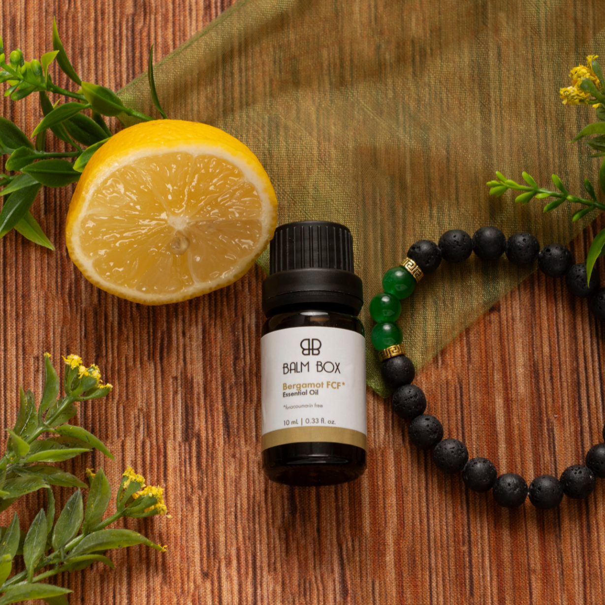 A bottle of Premium Soothing Skin & Lips, a sliced lemon, a black beaded bracelet with green accents, and green leaves rest on a wooden surface with a green cloth—an uplifting gift box for cancer care.