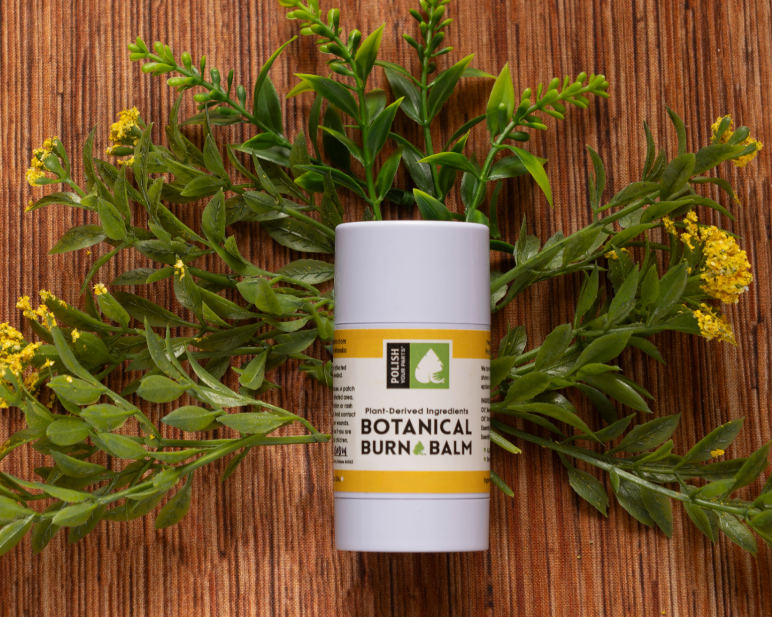 A tube of Premium Soothing Skin & Lips with a yellow label rests on a wooden surface amid green leaves and yellow flowers—a calming essential for any self-care routine, especially for cancer patients.