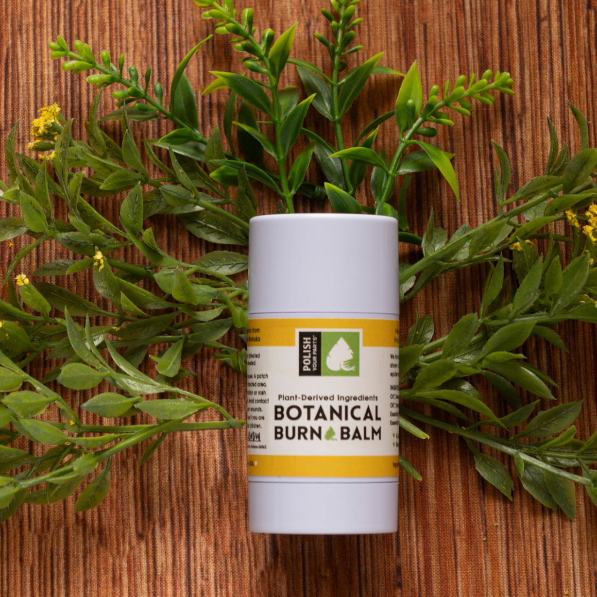 A tube of Premium Soothing Skin & Lips with a yellow label rests on a wooden surface amid green leaves and yellow flowers—a calming essential for any self-care routine, especially for cancer patients.