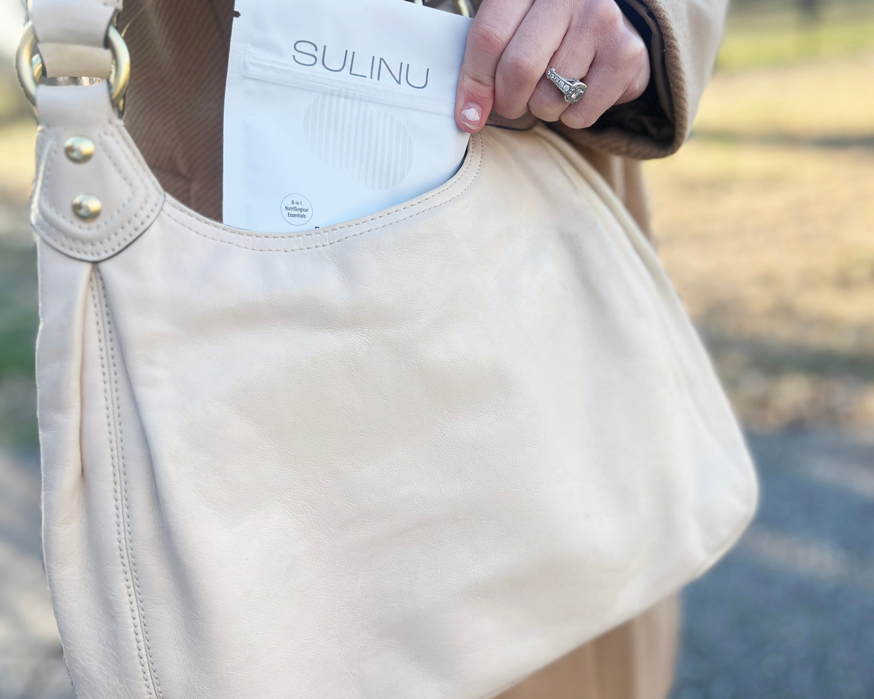 A person in a tan coat places a white Travel Packs-Set of 5-Single Serving Before + After Vitals vitamin powder packet into a light beige purse’s front pocket outdoors, with blurred greenery and pavement in the background.