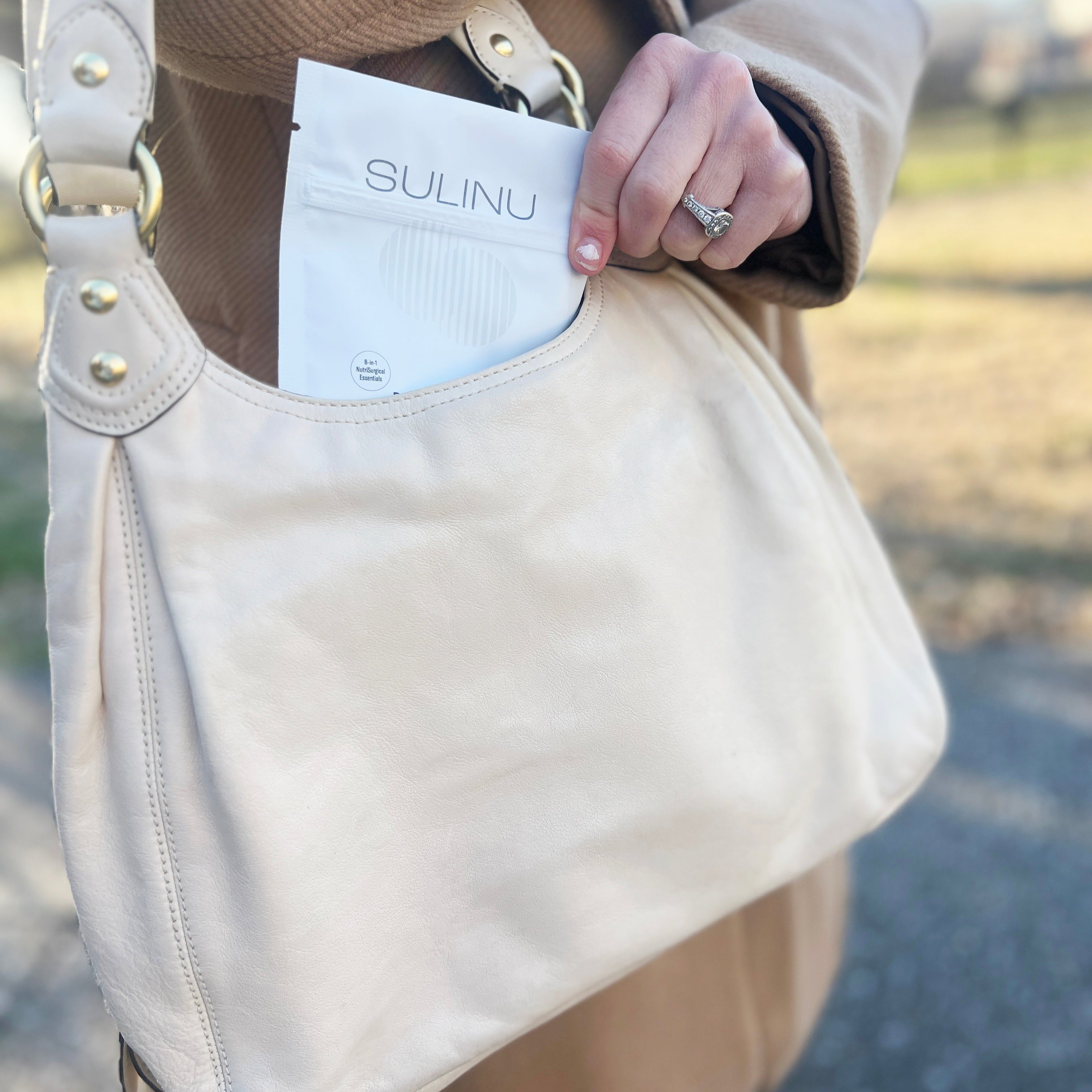 A person in a tan coat places a white Travel Packs-Set of 5-Single Serving Before + After Vitals vitamin powder packet into a light beige purse’s front pocket outdoors, with blurred greenery and pavement in the background.
