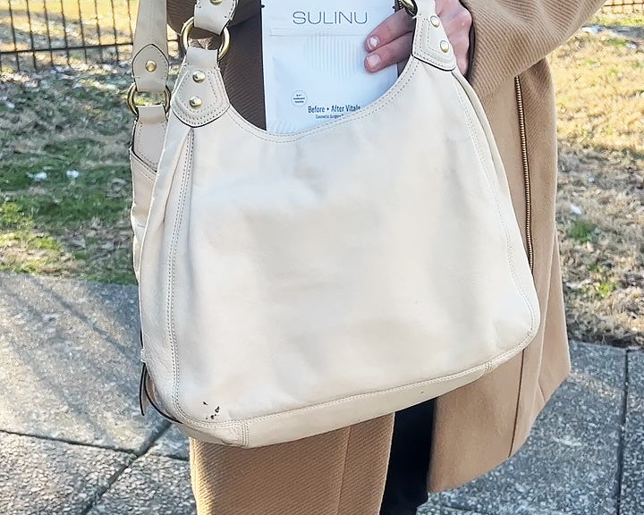 A person in a tan coat and black pants holds a beige handbag with a document labeled SULINU partly visible, possibly carrying Travel Packs- Set of 5- Single Serving Before + After Vitals to support wound healing after surgery.