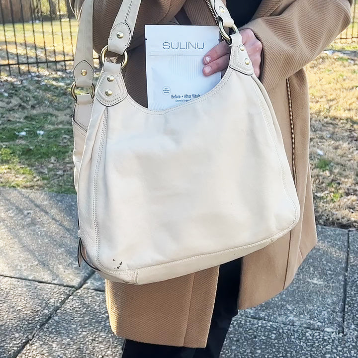 A person in a tan coat and black pants holds a beige handbag with a document labeled SULINU partly visible, possibly carrying Travel Packs- Set of 5- Single Serving Before + After Vitals to support wound healing after surgery.