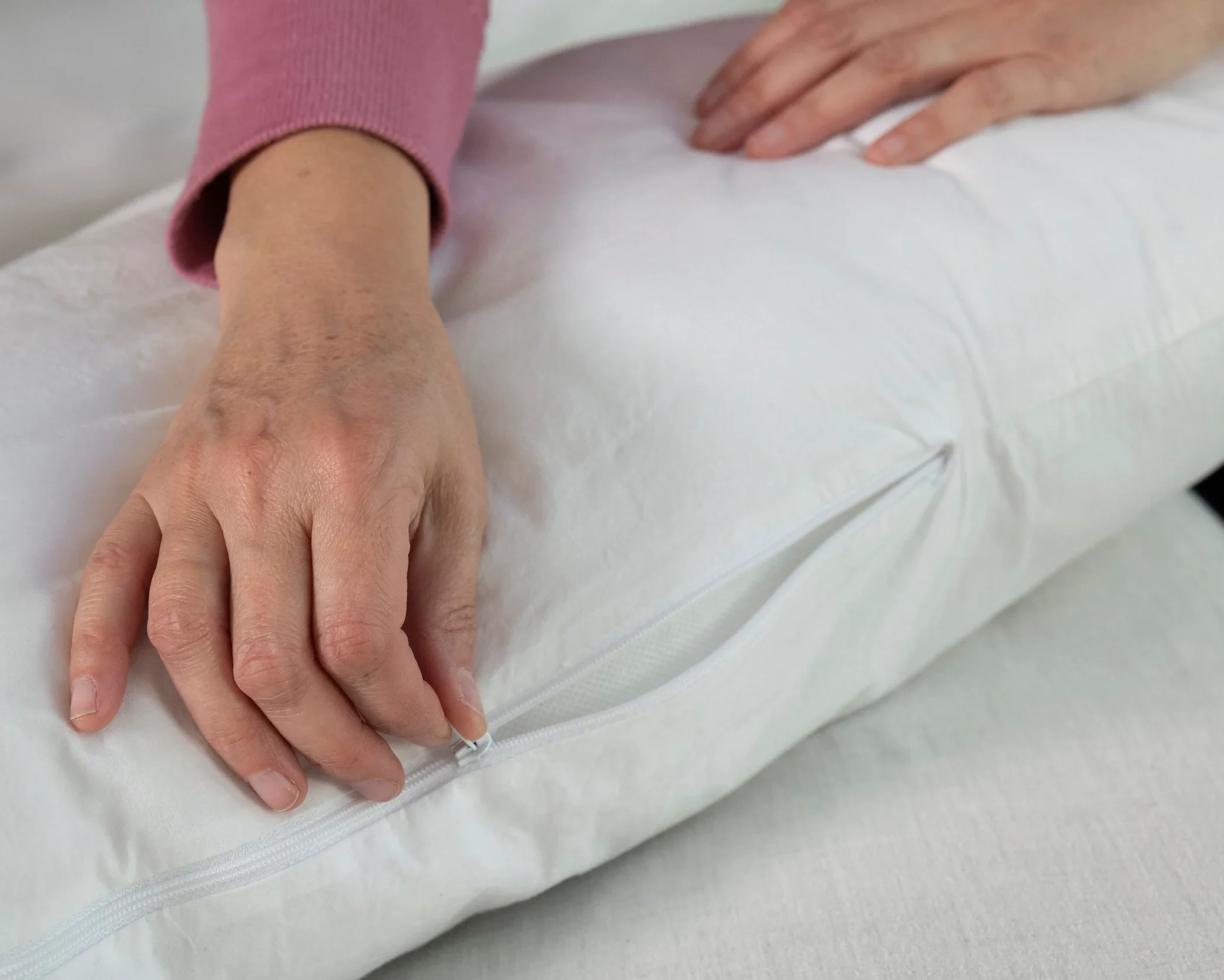 A person in a long-sleeved pink top unzips the washable slipcover of a white Two Side Pillow (sold separately), holding it gently on a light-colored surface and revealing the pillow inside.