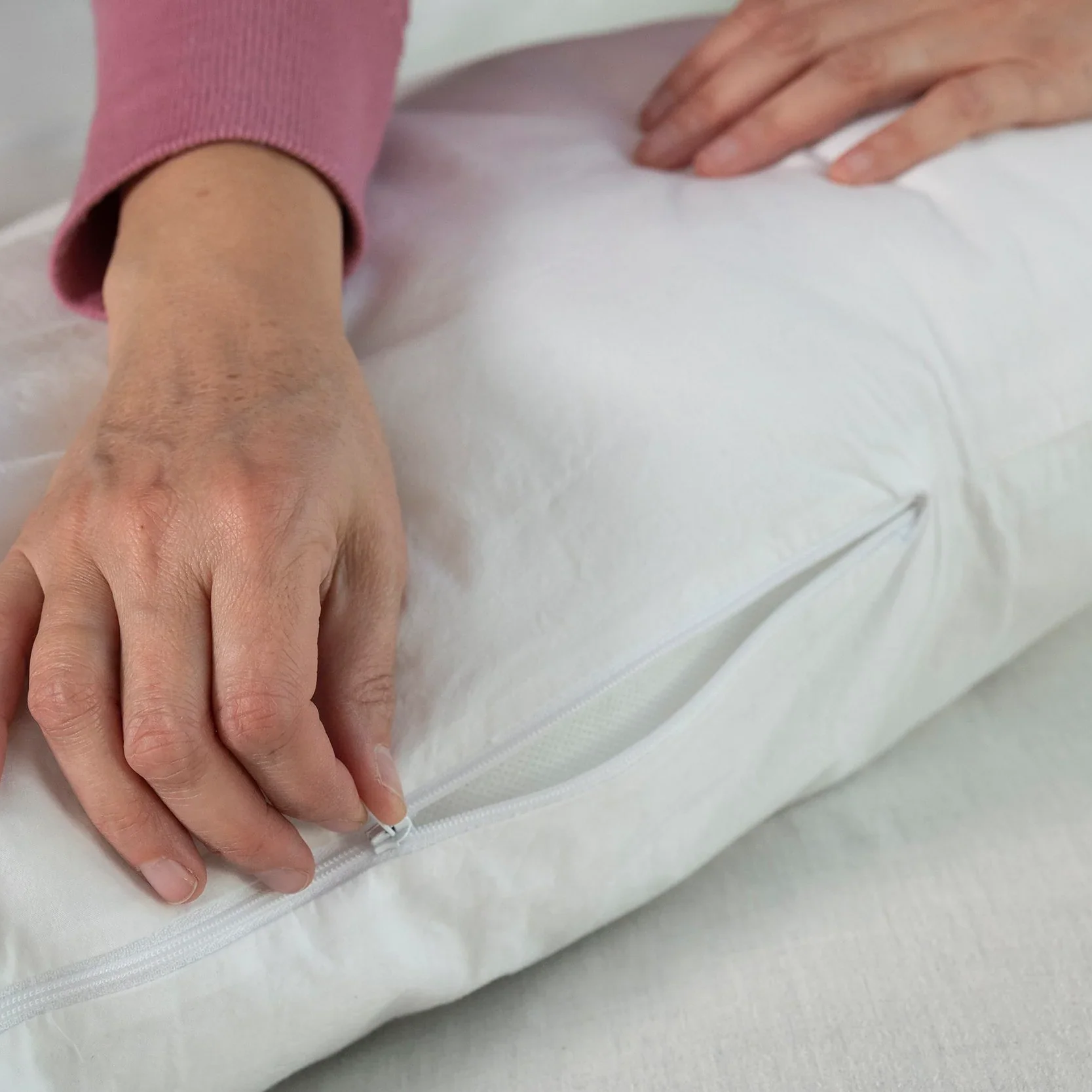 A person in a long-sleeved pink top unzips the washable slipcover of a white Two Side Pillow (sold separately), holding it gently on a light-colored surface and revealing the pillow inside.
