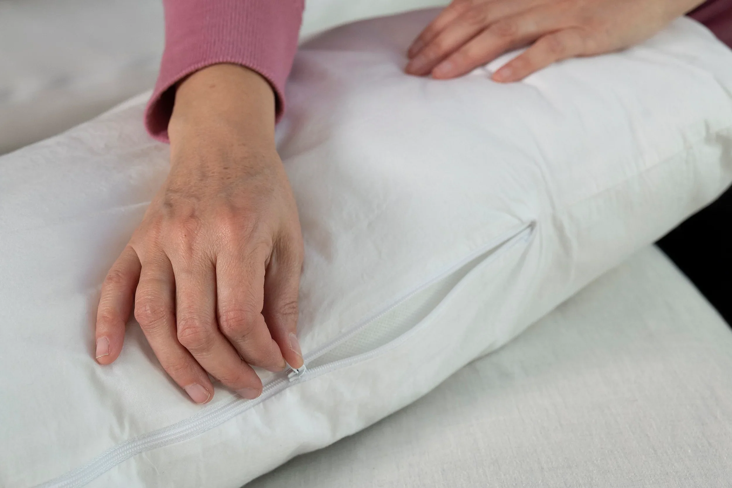 A person in a long-sleeved pink top unzips the washable slipcover of a white Two Side Pillow (sold separately), holding it gently on a light-colored surface and revealing the pillow inside.