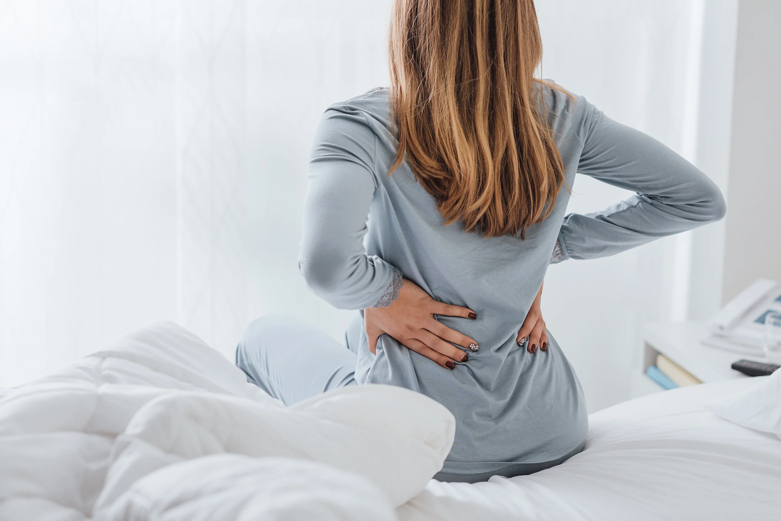 A woman clutches her lower back after waking up in pain. The Sleep Again Pillow System provides relief for anyone suffering from back pain, including chronic conditions or acute injuries.