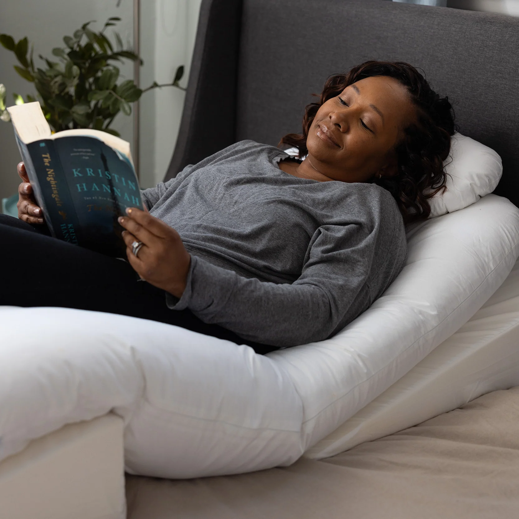 A woman relaxes on a bed, reading with a smile while supported by the white Sleep Again Pillow System Bundle. The softly lit room features a plant and a custom fitted sheet in the background.