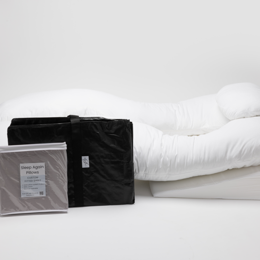 A Sleep Again Pillow System Bundle, featuring a white body pillow and wedge pillow, sits on a white background with a folded black travel bag and packaged pillowcases labeled Sleep Again Pillows in front.