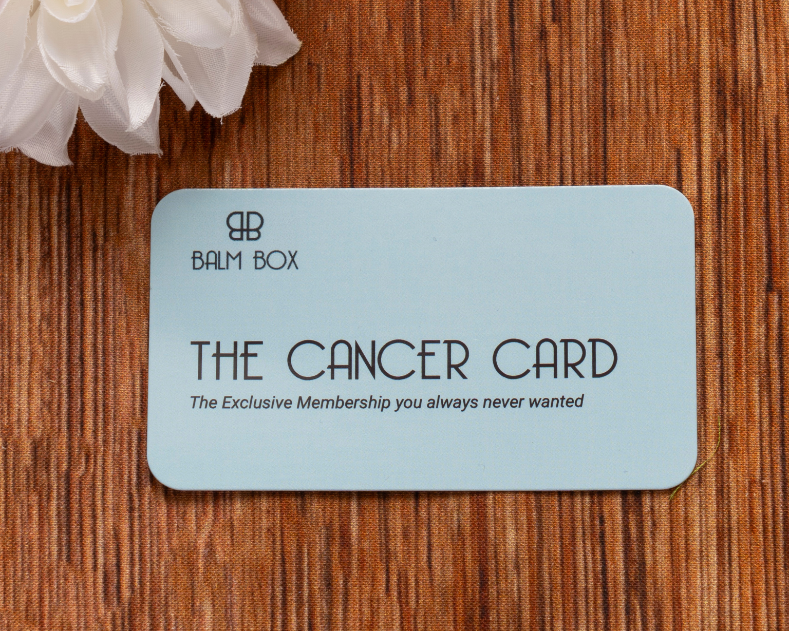 A light blue card labeled Sweetest Rest & Recovery Box lies on a brown textured surface next to a white flower. The card reads, The Exclusive Membership you always never wanted—a thoughtful self care for cancer patients gift.