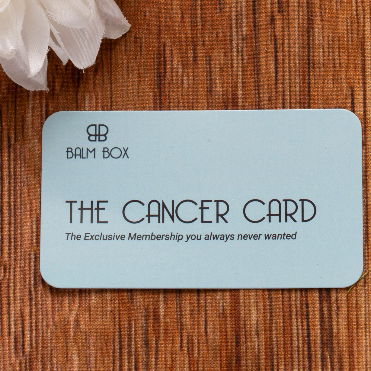 A light blue card labeled Sweetest Rest & Recovery Box lies on a brown textured surface next to a white flower. The card reads, The Exclusive Membership you always never wanted—a thoughtful self care for cancer patients gift.