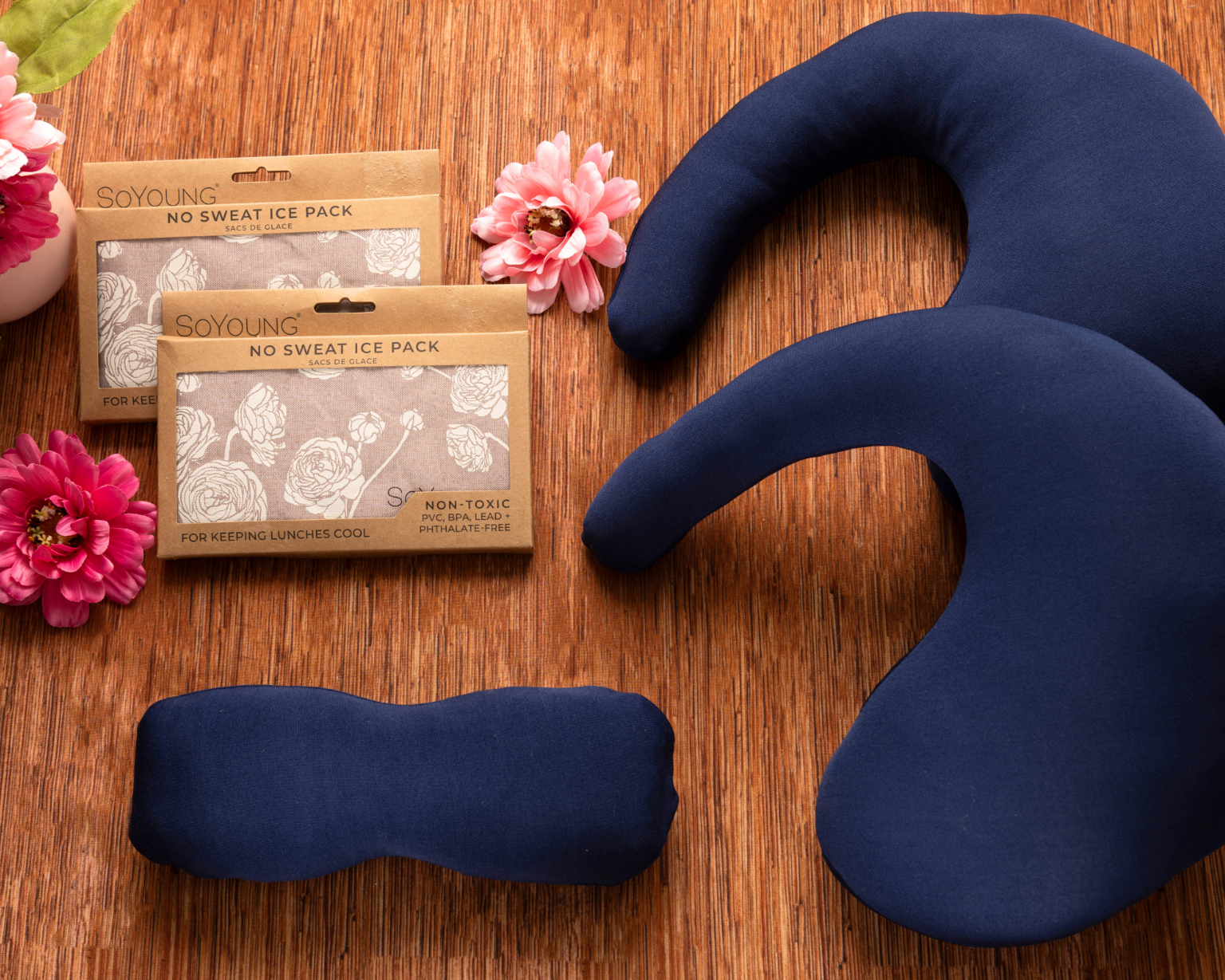 The Best Mastectomy Care Package features navy blue fabric therapy packs and two brown No Sweat Ice Pack boxes, arranged on a wooden surface with three pink artificial flowers, creating a thoughtful gift for recovery.