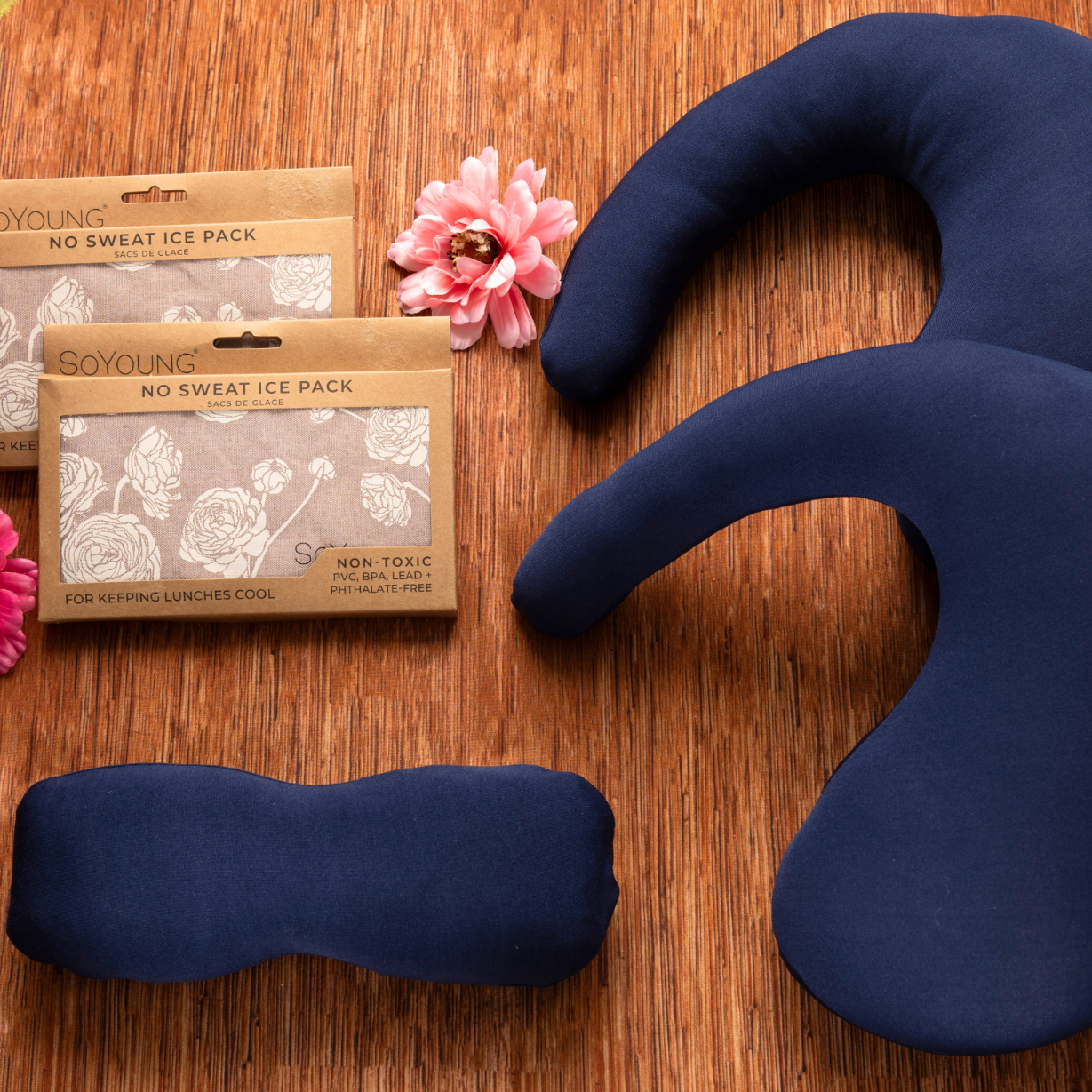 The Best Mastectomy Care Package features navy blue fabric therapy packs and two brown No Sweat Ice Pack boxes, arranged on a wooden surface with three pink artificial flowers, creating a thoughtful gift for recovery.