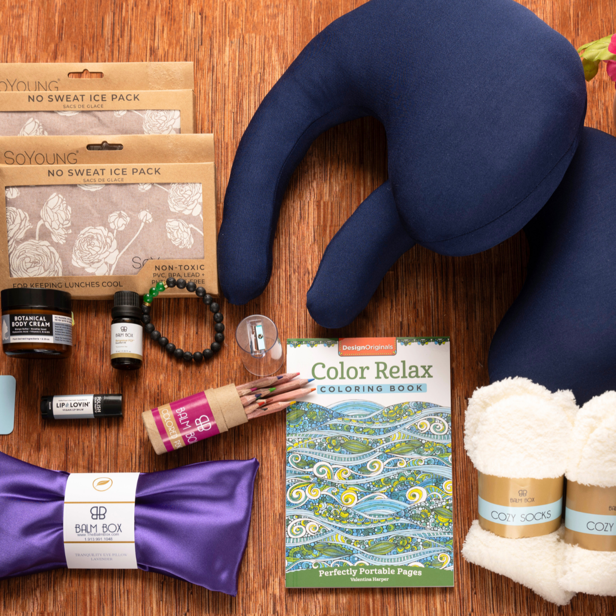 The Best Mastectomy Care Package sits on a wooden surface, featuring a post-mastectomy pillow, cozy socks, coloring book with pencils, ice packs, lotion, eye mask, bracelet, membership card, and pink flowers.