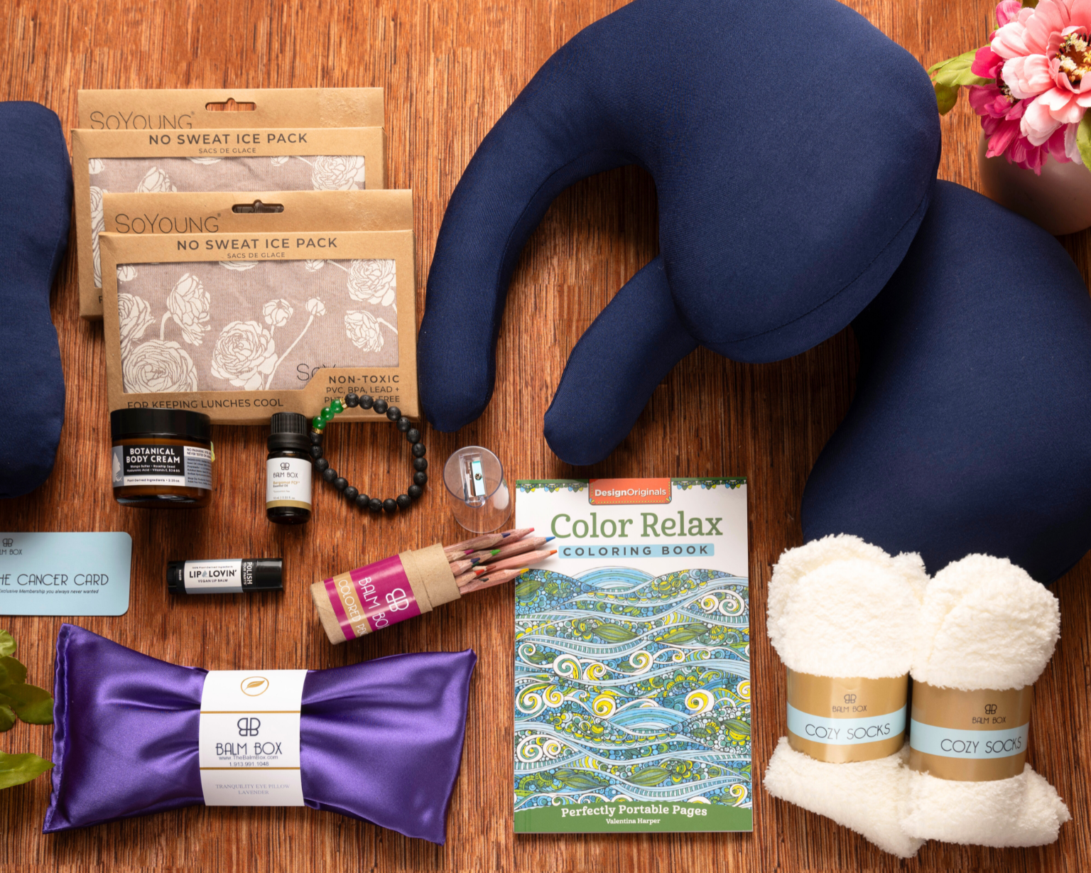 A flat lay of The Best Mastectomy Care Package features a neck pillow, ice packs, cozy socks, colored pencils, coloring book, satin eye mask, flowers, bracelet, candle, and balm—thoughtfully assembled for breast cancer or mastectomy recovery.