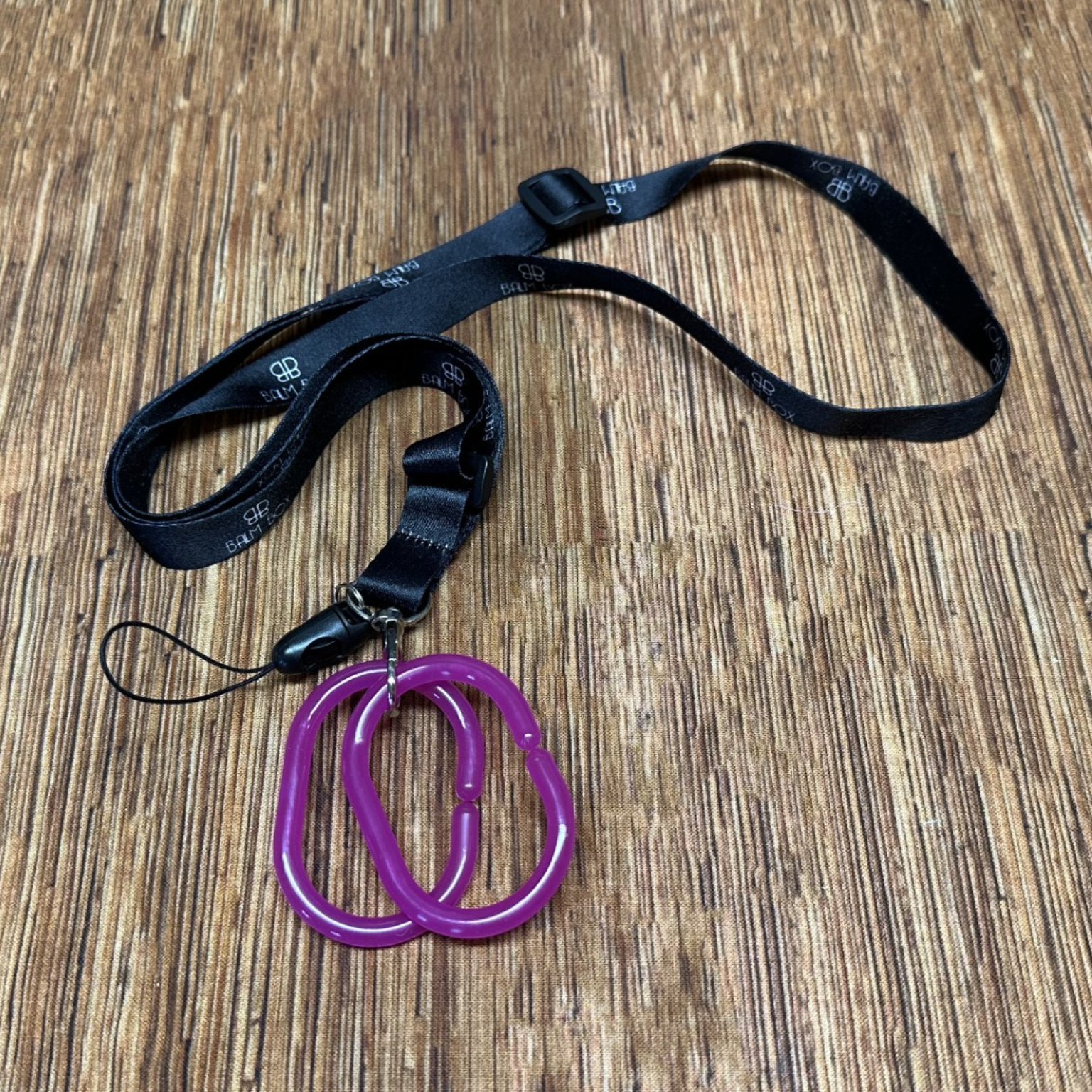 The Surgical Drain Holder features a black lanyard with white text and two linked purple carabiners on a small strap, shown on a wooden surface for practical at-home drain management.