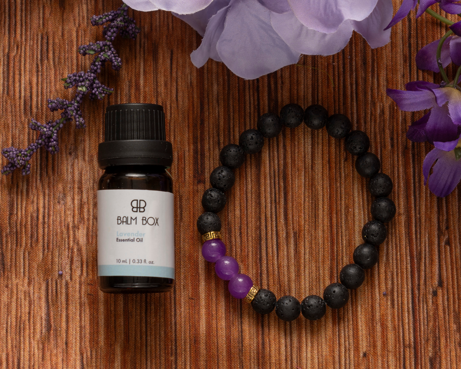 A small bottle of Sweetest Rest & Recovery Box essential oil sits beside a black beaded bracelet with three purple beads on a striped brown surface, surrounded by purple flowers—a thoughtful self-care gift for cancer patients.